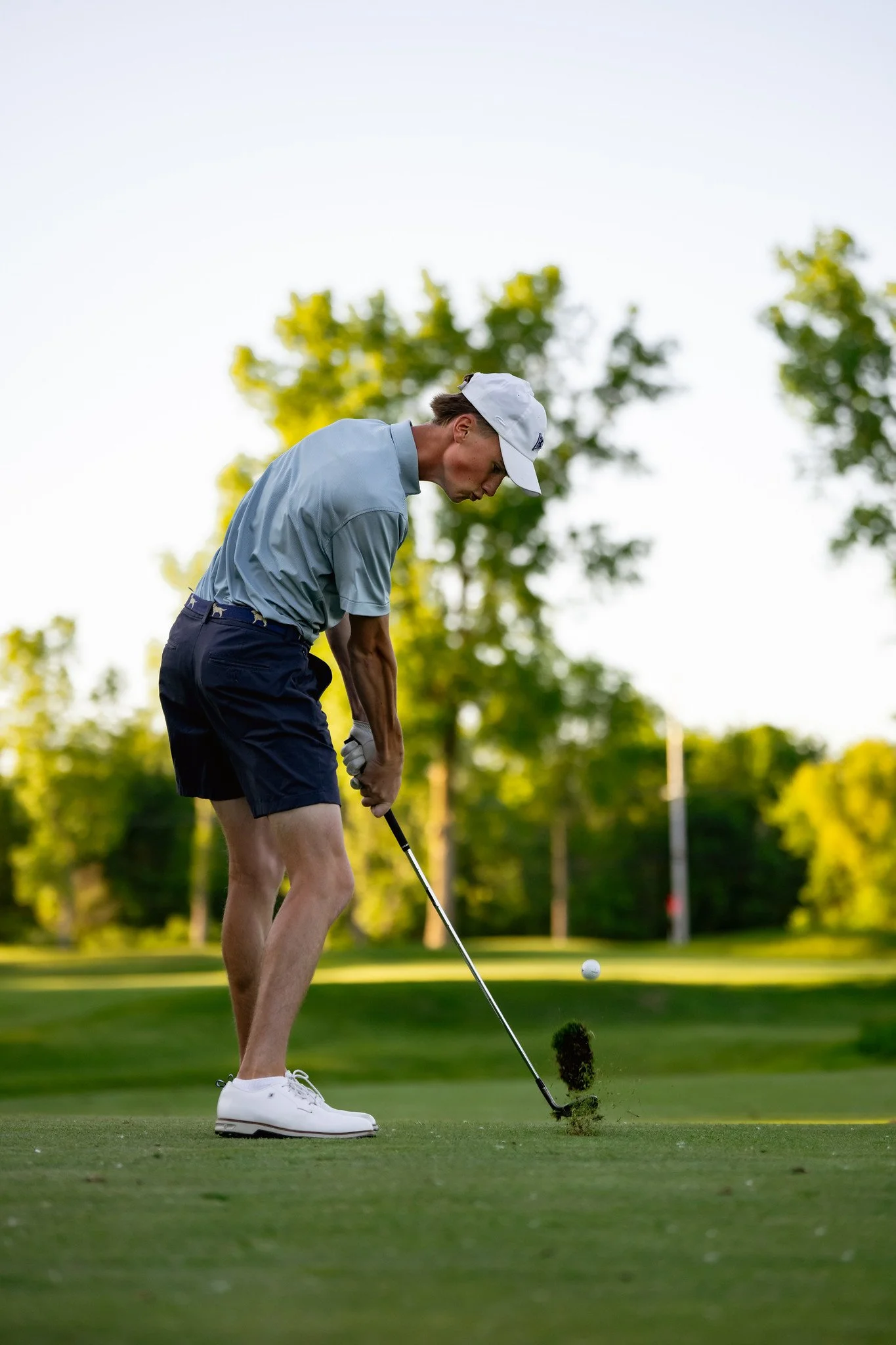 A person in golf attire, including a white cap, light blue shirt, black shorts, and white shoes, is preparing to hit a golf ball on a well-maintained golf course with trees and flags in the background.