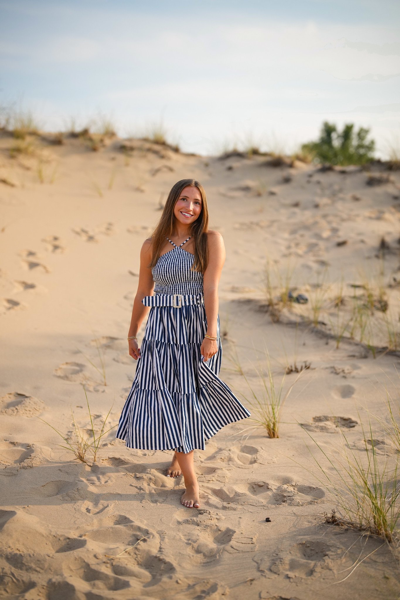 Woman in a blue and white striped dress walking barefoot on sandy beach dunes with grass and shrubs.