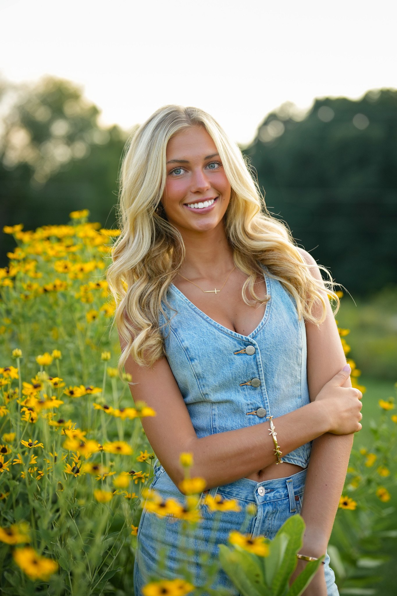 A young woman with long blonde hair and blue eyes, smiling and standing in a field of yellow flowers with green trees in the background, during daytime.