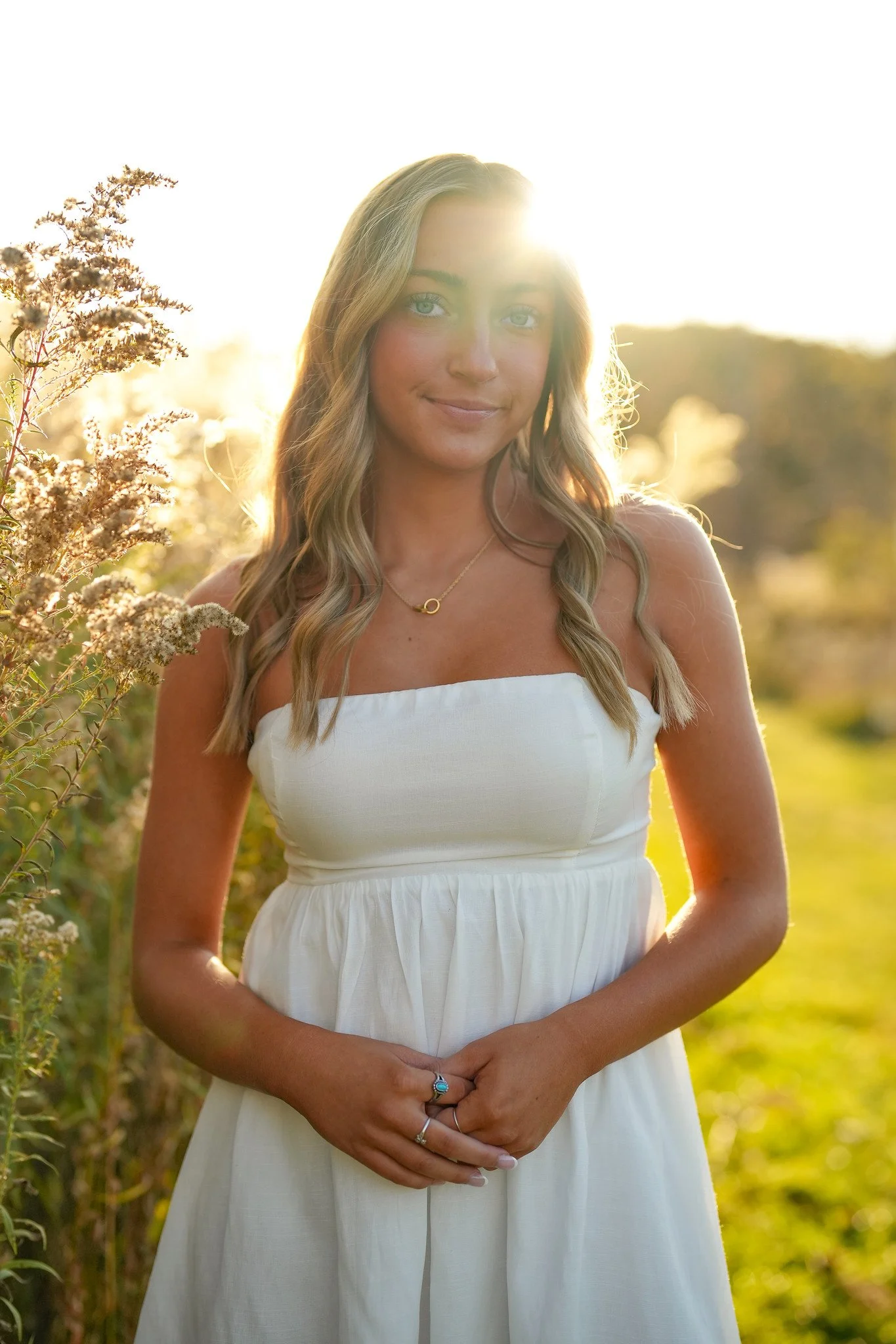 A woman with long wavy blonde hair and light blue eyes standing outdoors during sunset, wearing a white strapless dress and jewelry, with gold and silver rings, surrounded by tall plants and golden sunlight.