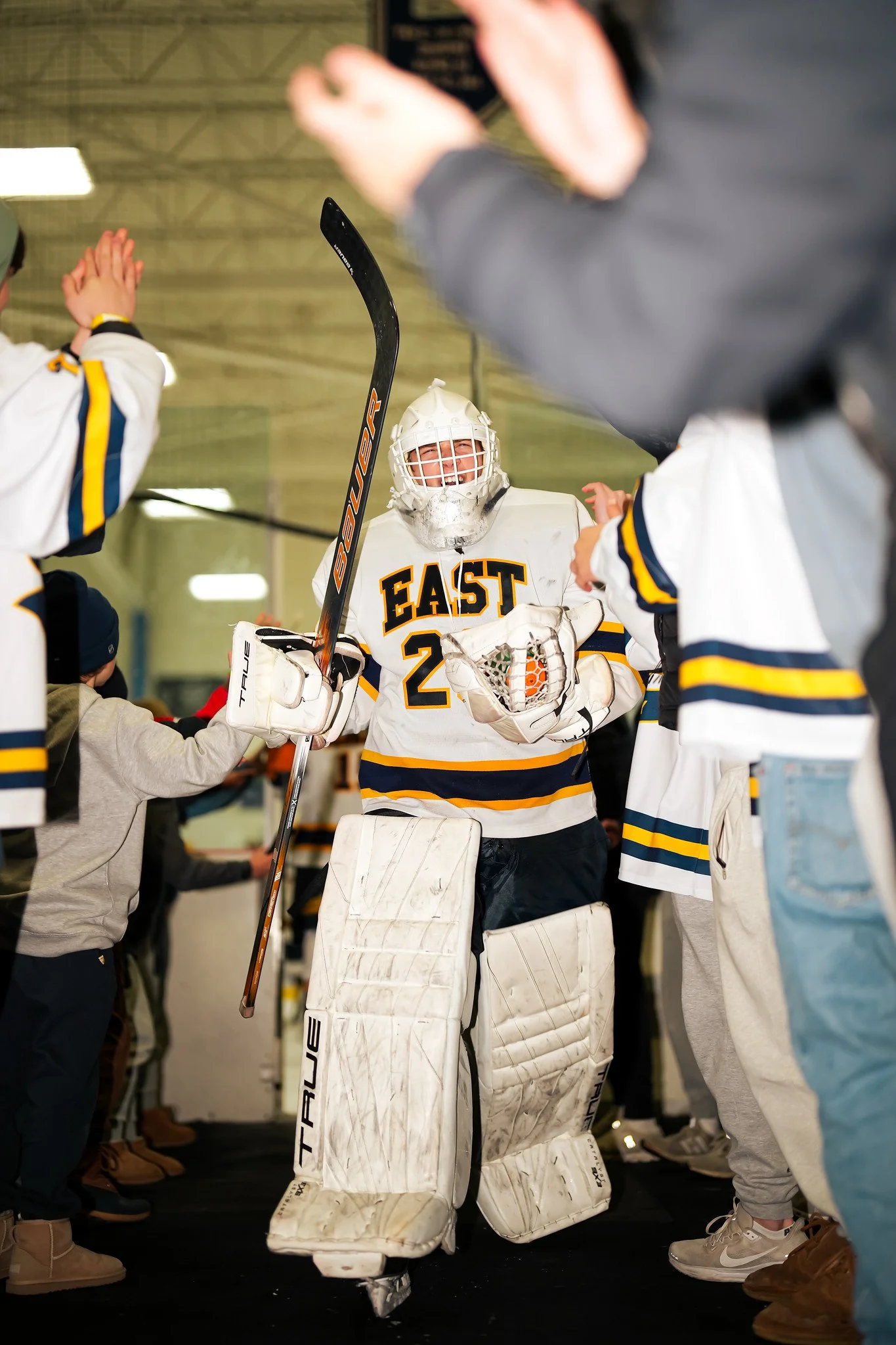 A hockey goalie in a white uniform with the word 'EAST' on the chest, wearing a helmet and protective gear, standing among other players and fans in an indoor rink.