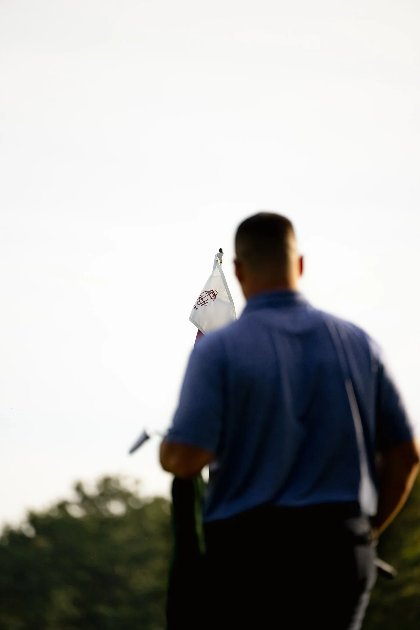 Person with short hair wearing a blue shirt holds a flag with a logo on it, standing outdoors during daytime with trees in the background.