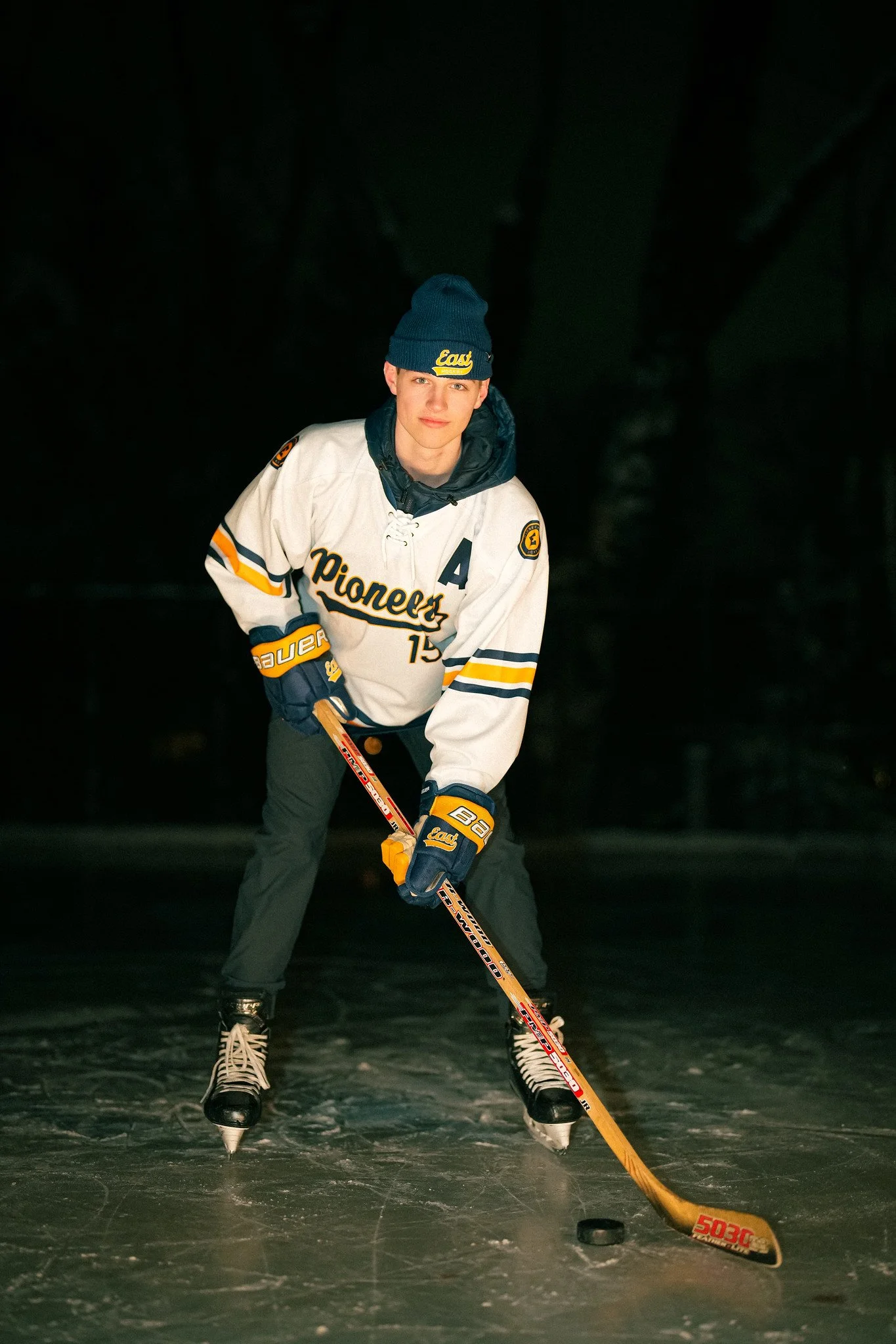 A young man in ice hockey gear, including skates, gloves, and a jersey that says 'Pioneers', is holding a hockey stick and playing on an ice rink at night. He is wearing a blue beanie with yellow writing and is in a ready stance, facing the camera.