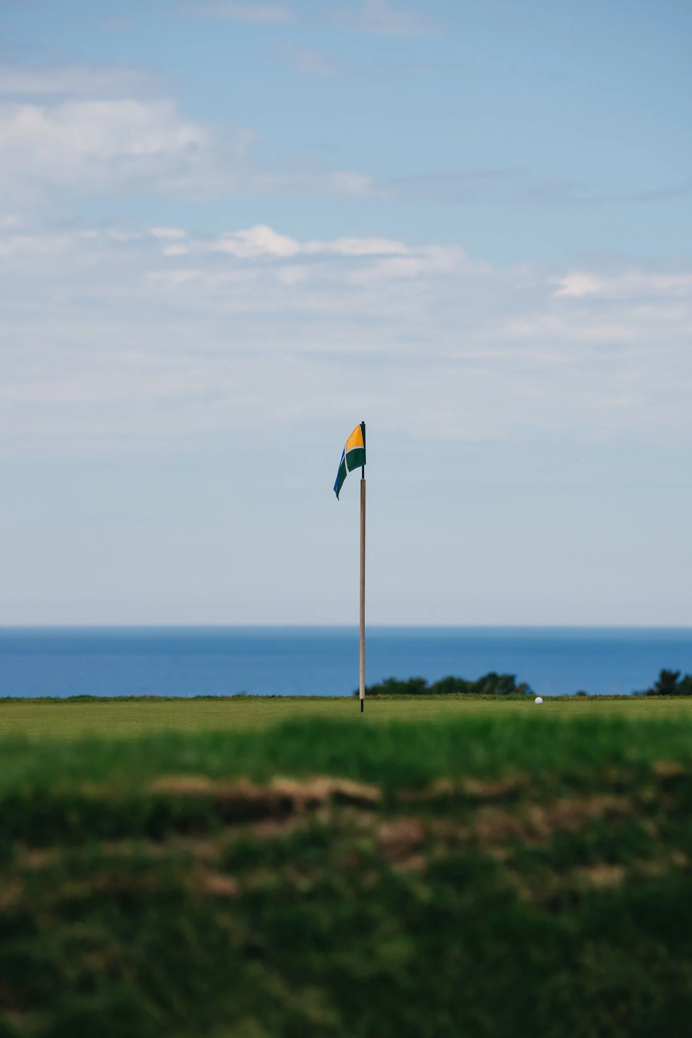A golf course with a flagstick and a golf ball below the flag, overlooking the ocean near the horizon with blue sky and some clouds.