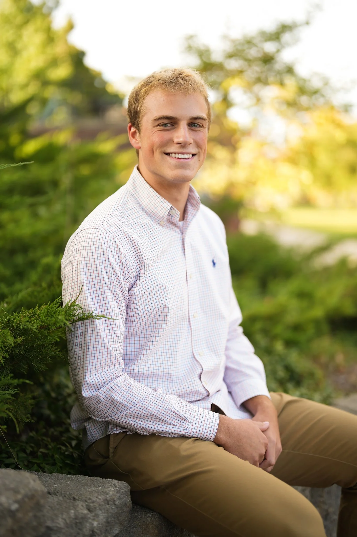 A young man with light brown hair, blue eyes, and fair skin sitting outdoors on a stone surface, smiling at the camera. He is wearing a white checkered shirt and tan pants, with a blurred background of green trees and bright sunlight.