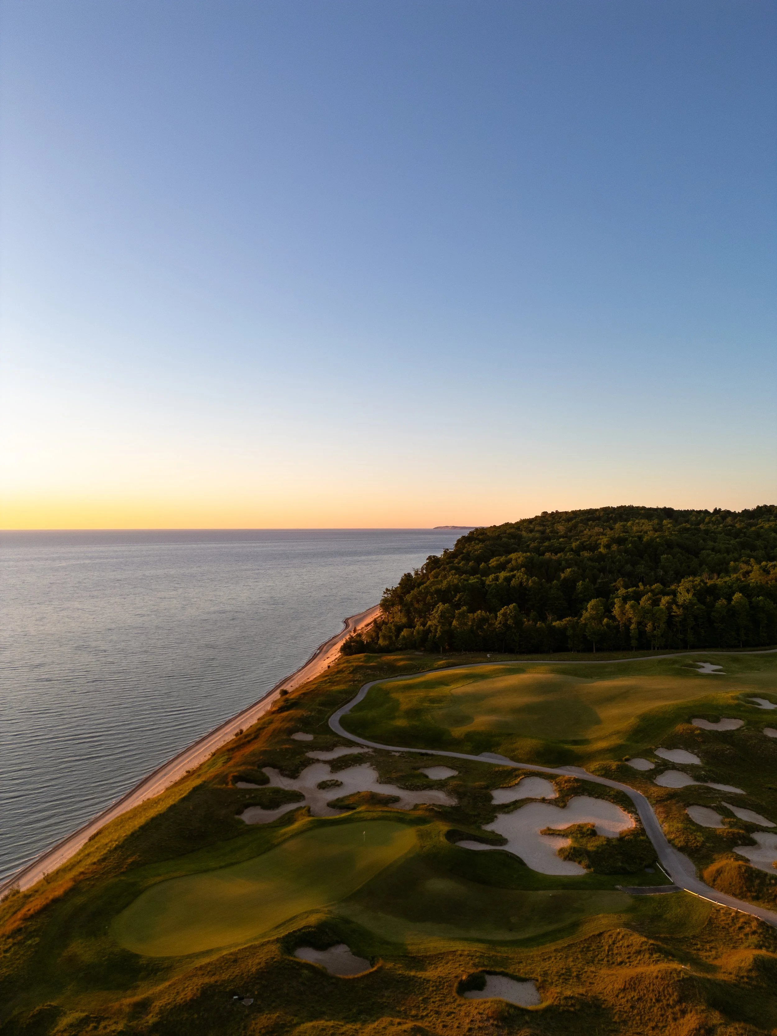 Aerial view of a golf course along a coastline during sunset, with a dense forested area, sand traps, green fairways, and a body of water.