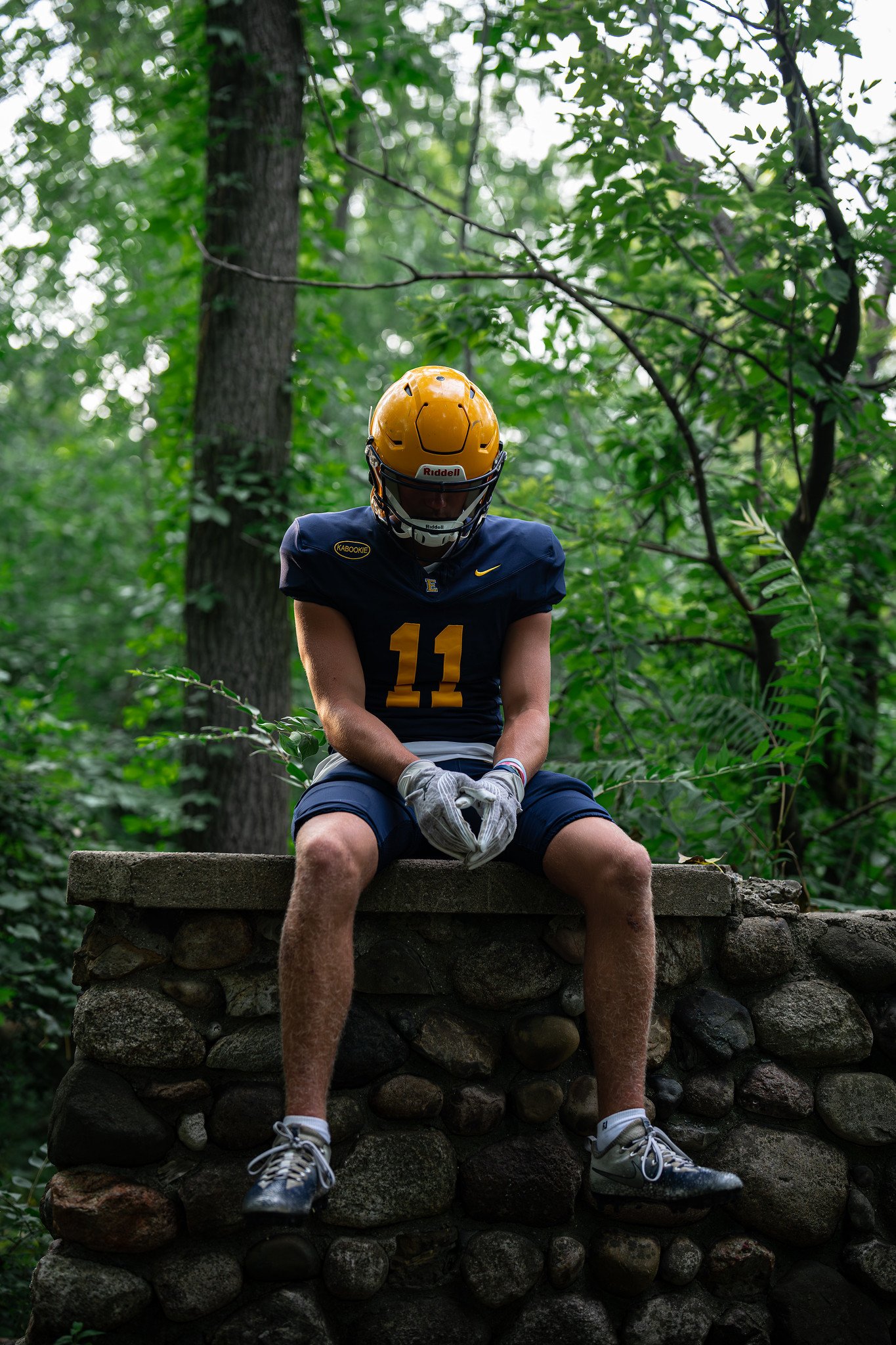 A football player in uniform with the number 11, wearing a yellow helmet, sitting on a stone wall in a forest setting.