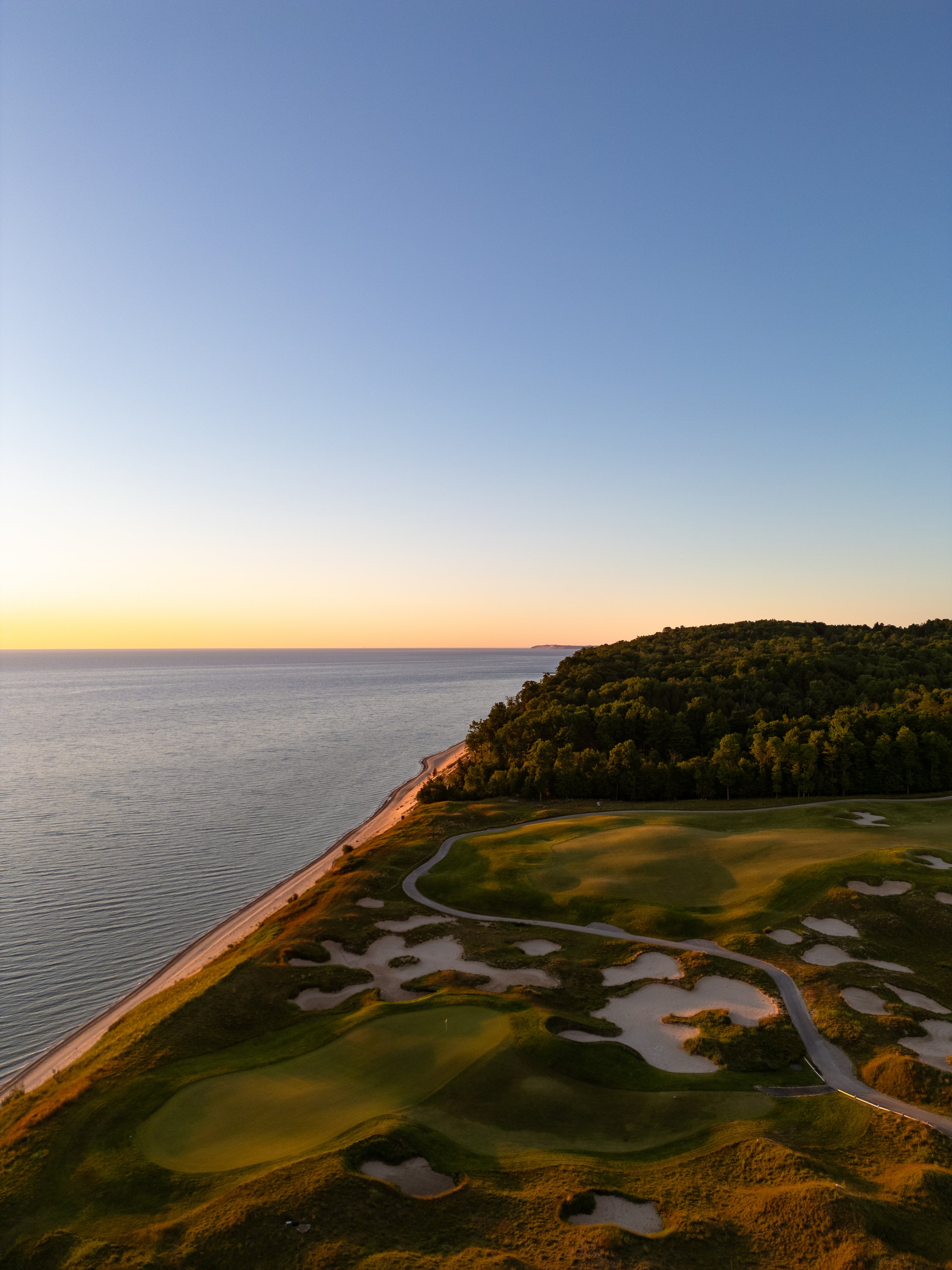 Aerial view of a golf course on a coastal hillside during sunset with a calm sea in the background and clear sky.