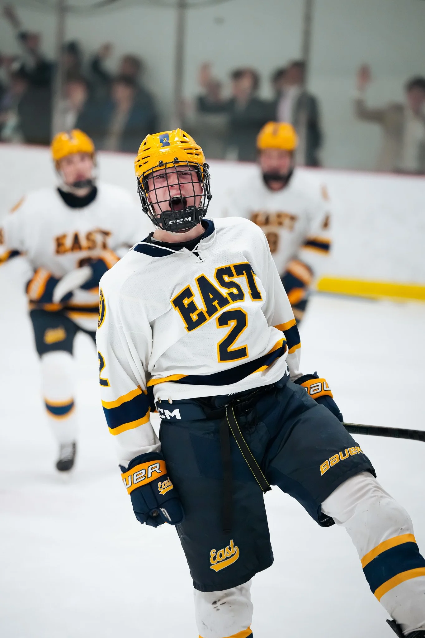 A young hockey player celebrating on the ice, wearing a white jersey with yellow and black accents, a yellow helmet, and black hockey pants. In the background, other teammates and spectators are visible.