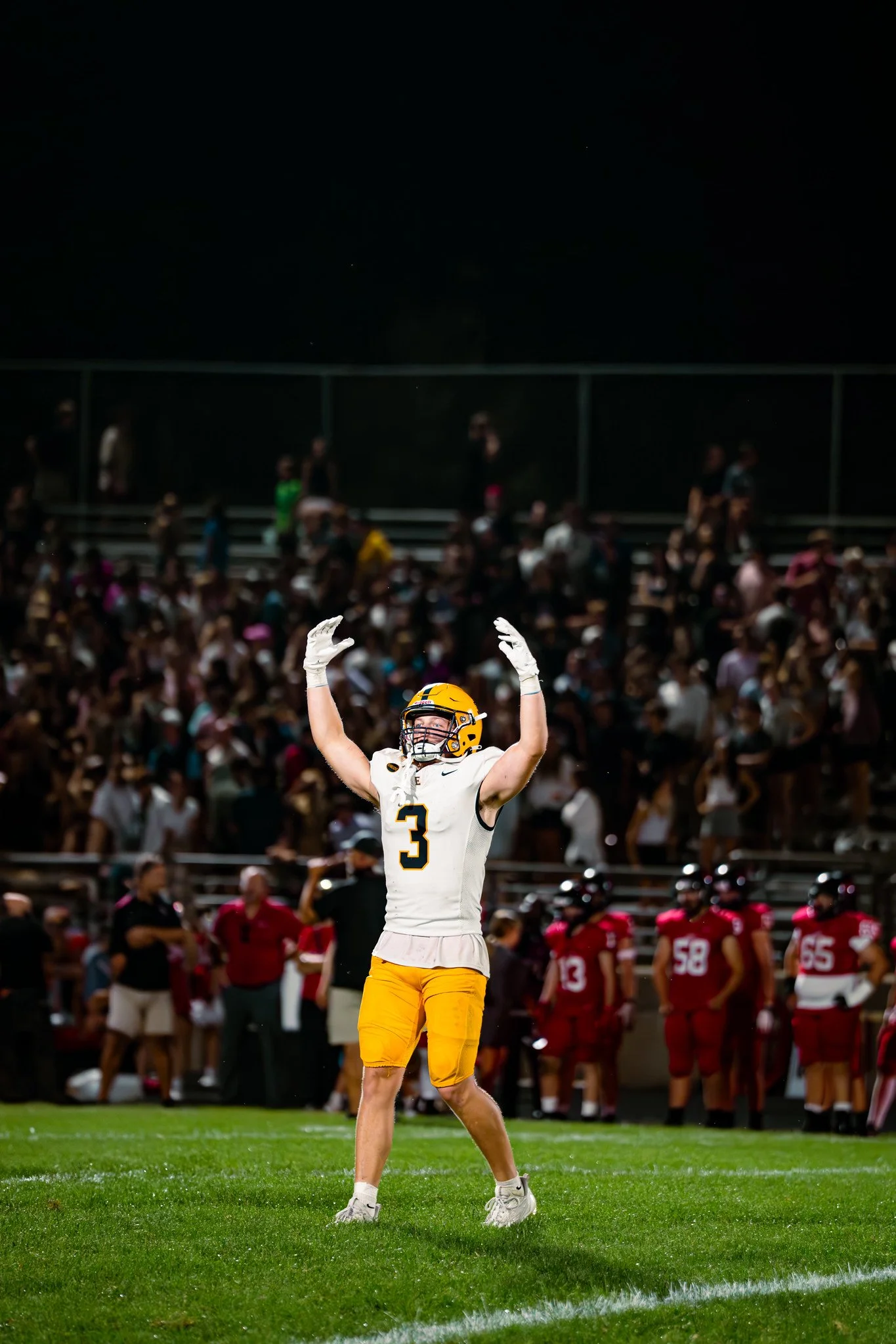 A football player wearing a white and yellow uniform, with the number 3 on his jersey, raising his hands during a game on a well-lit field with a crowd of spectators in the background.