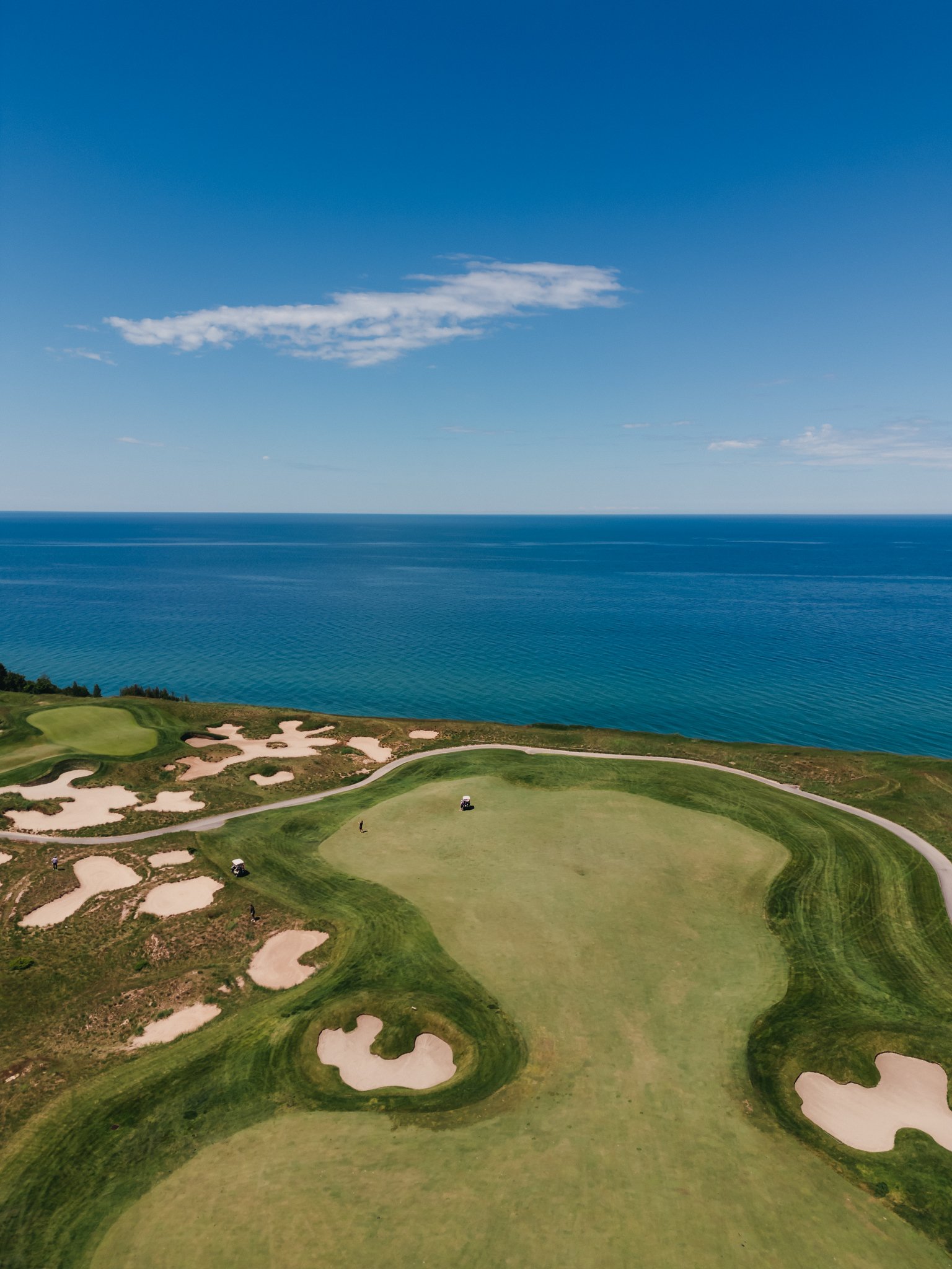 A golf course with green fairways, sand bunkers, and golf carts, near a large body of water under a blue sky with clouds.