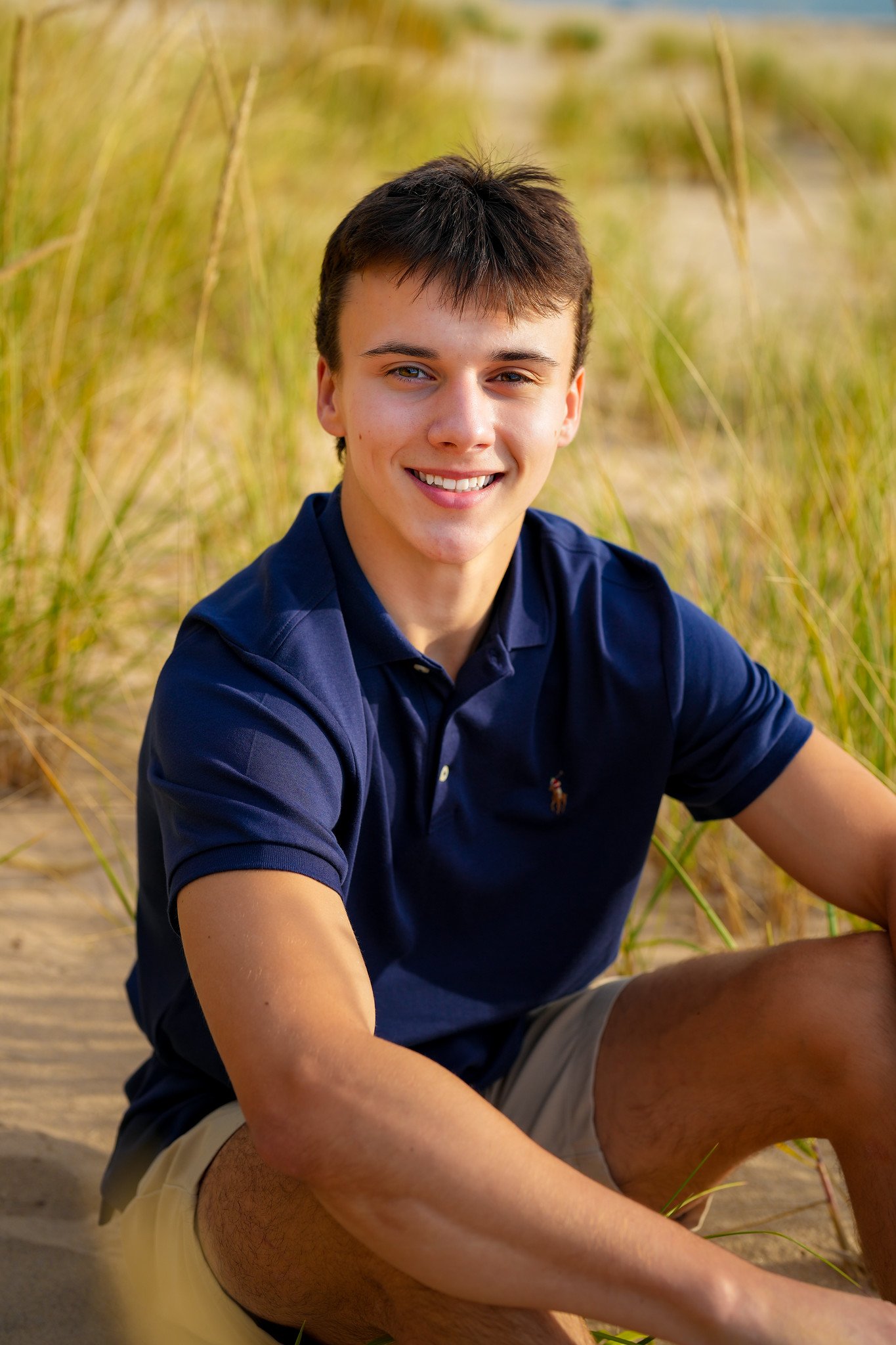 A young man with short dark hair smiling while sitting on a sandy beach in front of tall grass, wearing a navy blue polo shirt and beige shorts.