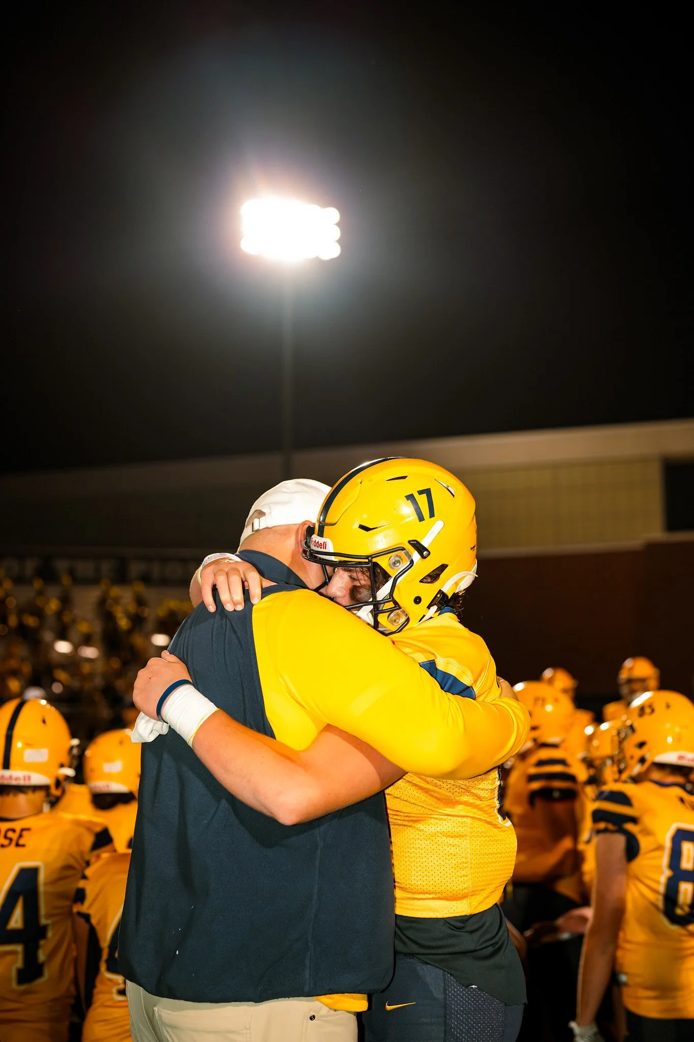 A football player and a coach hugging on the field at night during a game, with a stadium light shining brightly in the background and other players in yellow uniforms in the background.
