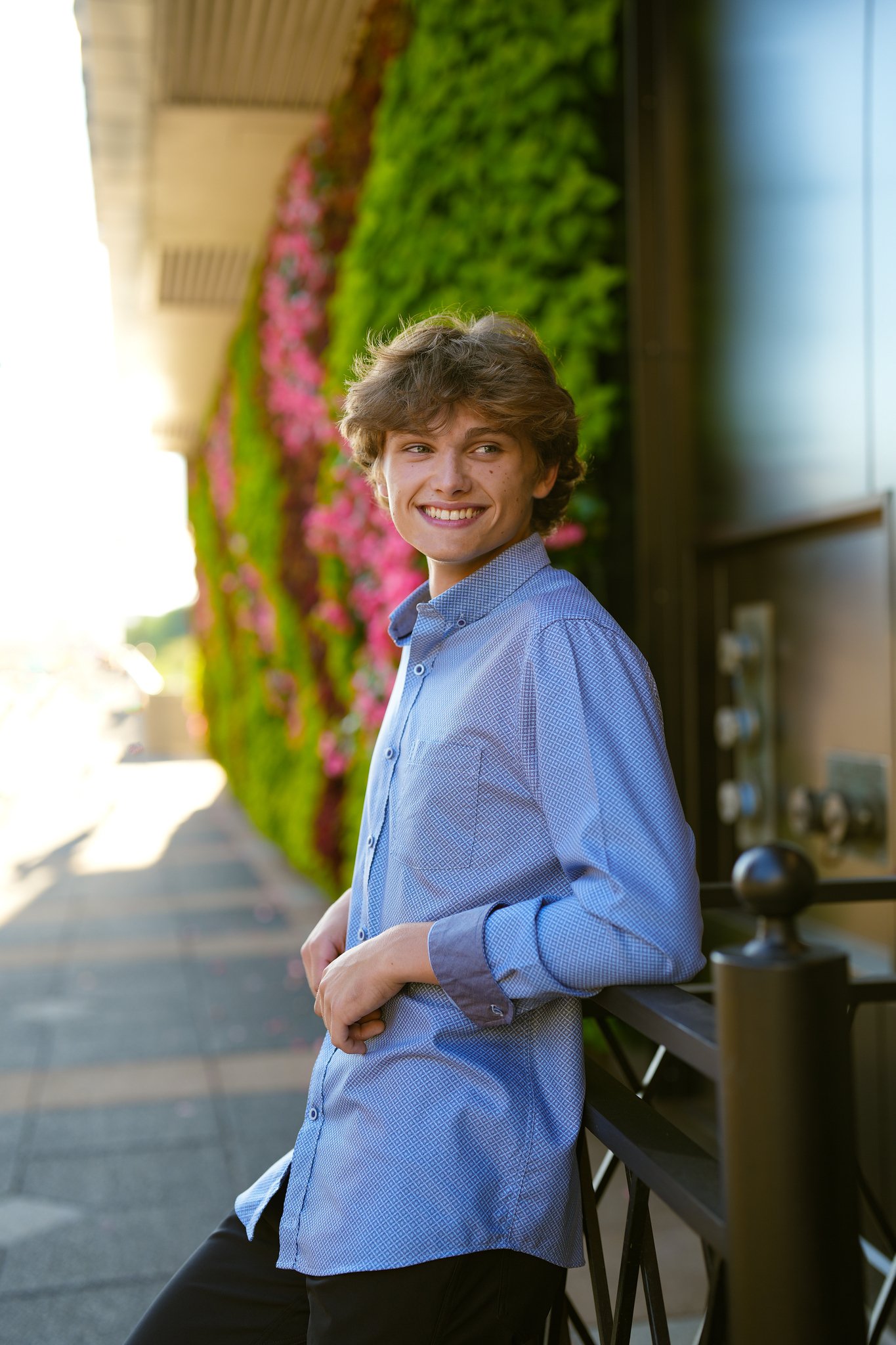 A young man with wavy brown hair and light skin smiling, wearing a blue dress shirt, leaning against a black railing outside with green and pink flowering plants in the background, during late afternoon.