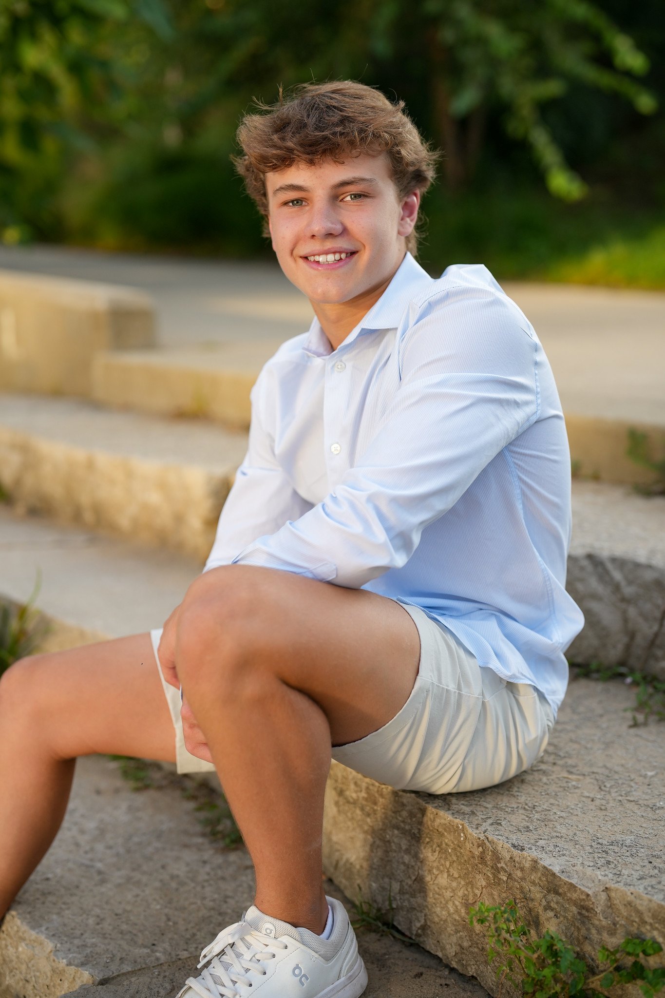 A young man sitting on a stone ledge outdoors, smiling, wearing a white shirt, beige shorts, and white sneakers, with greenery in the background.