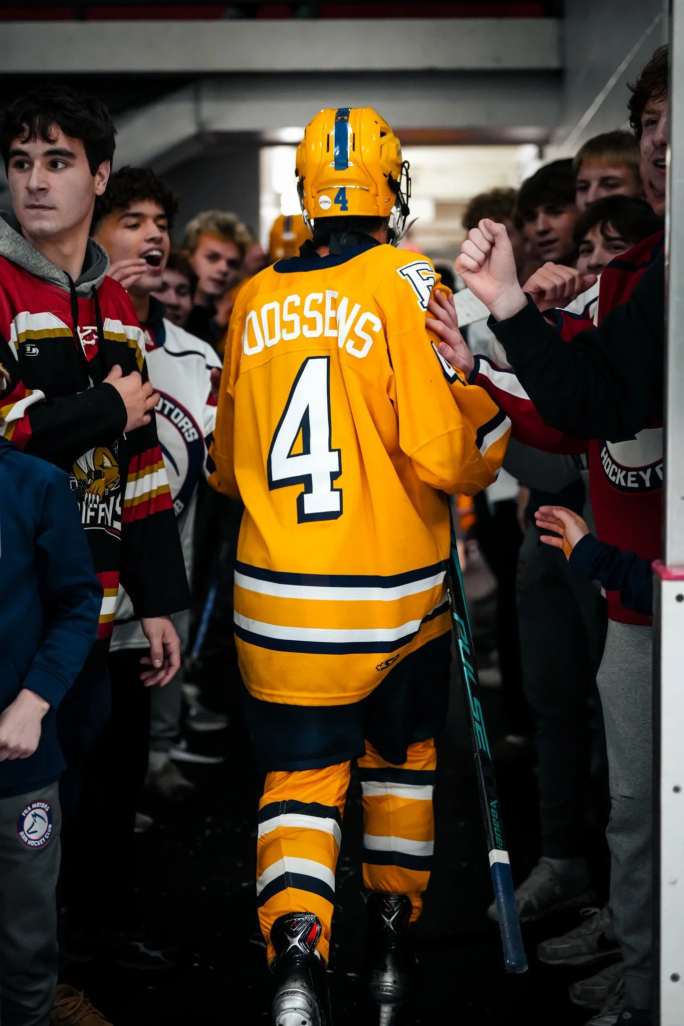 An ice hockey player in a yellow jersey with the name 'Dossevs' and the number 4, wearing a yellow helmet, is walking through a tunnel surrounded by fans who are reaching out to him, some wearing hockey jerseys.