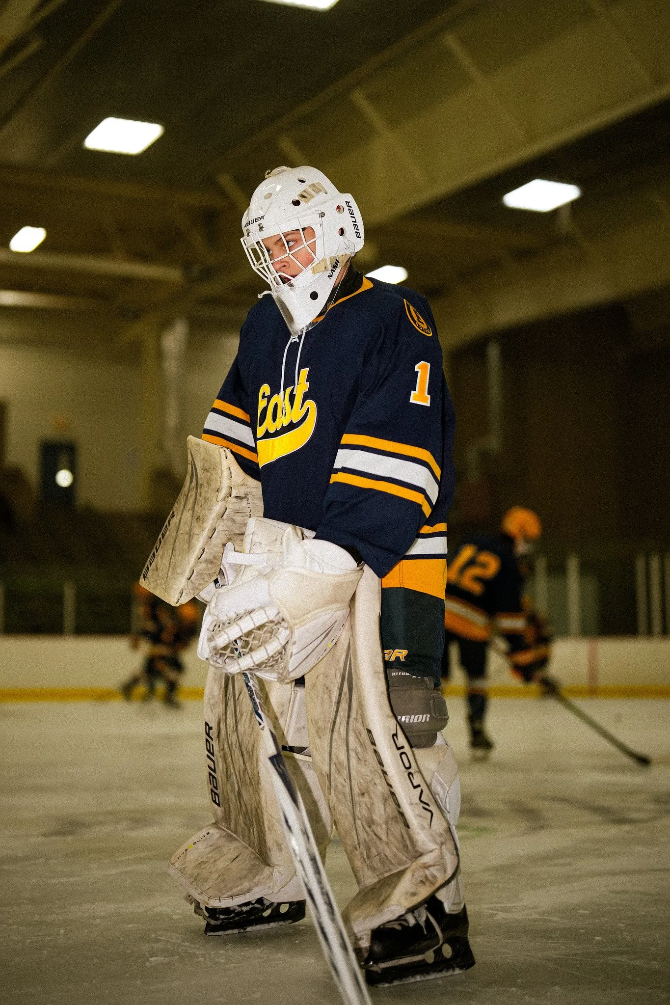 A hockey goalie wearing a white helmet and navy blue jersey with yellow accents, standing on the ice rink with a focused expression.