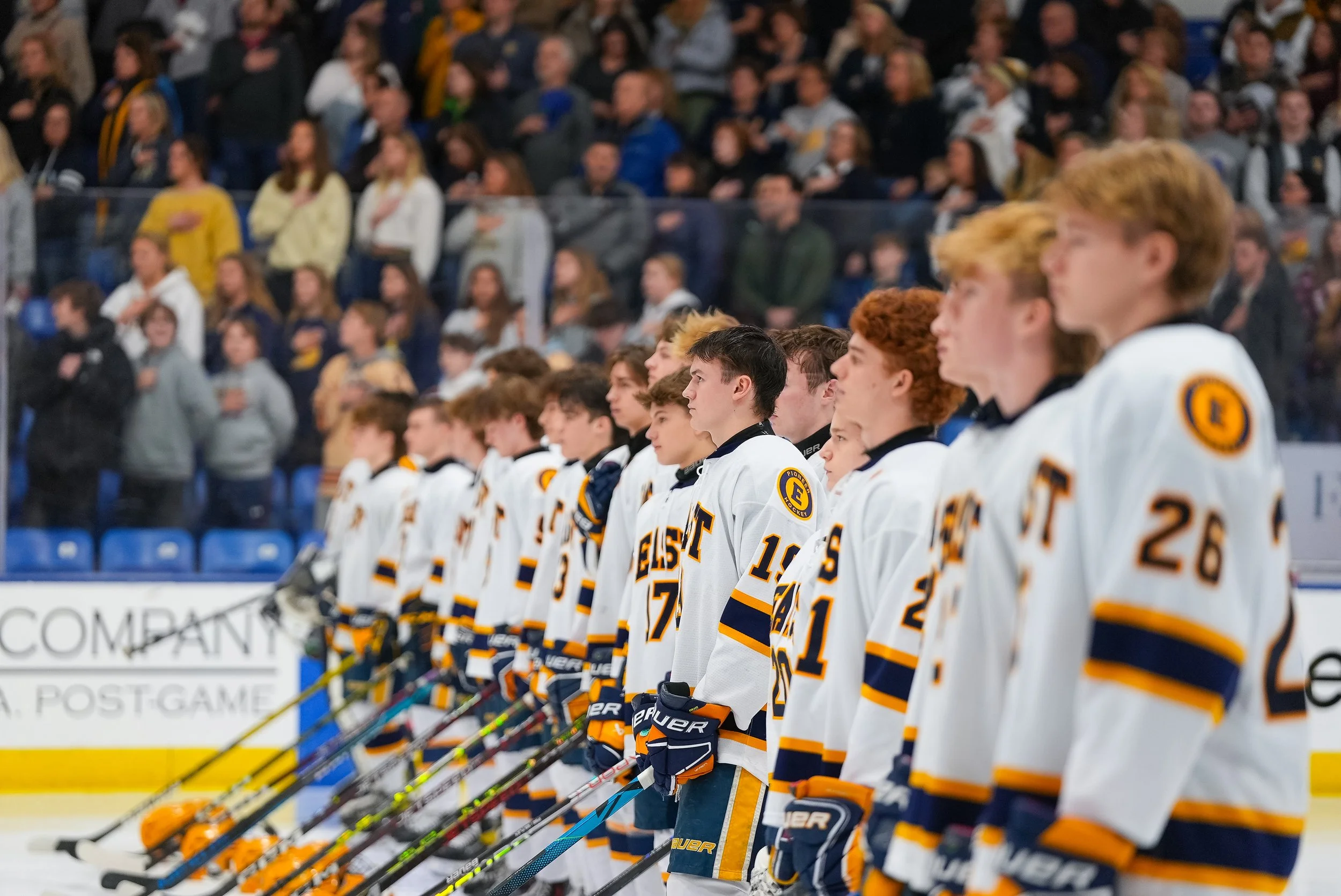 An ice hockey team in white jerseys with yellow and black accents standing in a line on the ice, facing forward, likely during the national anthem or a pre-game ceremony, with spectators in the background.