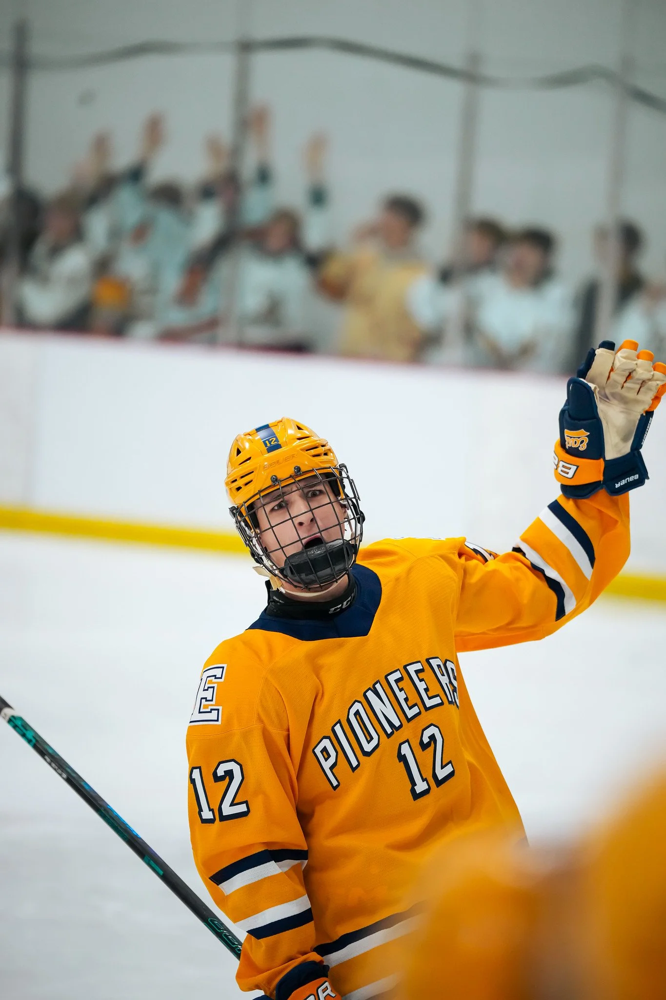 An ice hockey player in a yellow jersey with the number 12 on the back, wearing a yellow helmet and gloves, is raising a hand during a game.
