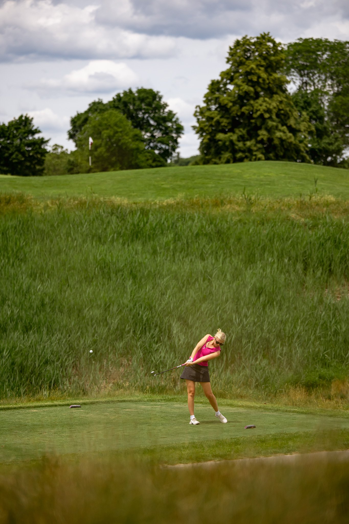 A woman teeing off on a golf course, wearing a pink top and skirt, with trees and clouds in the background.