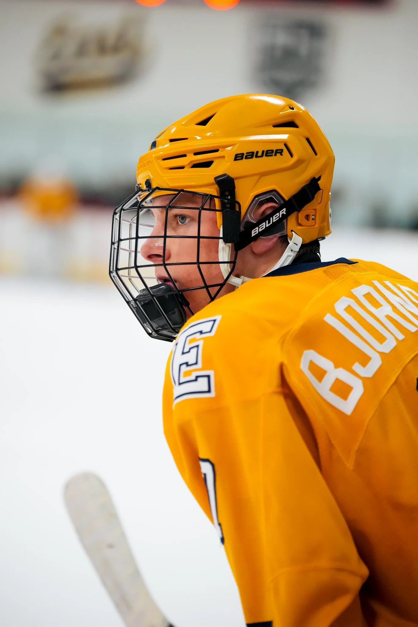 Close-up of a hockey player wearing a yellow helmet with a black cage and a yellow jersey, on an ice rink.