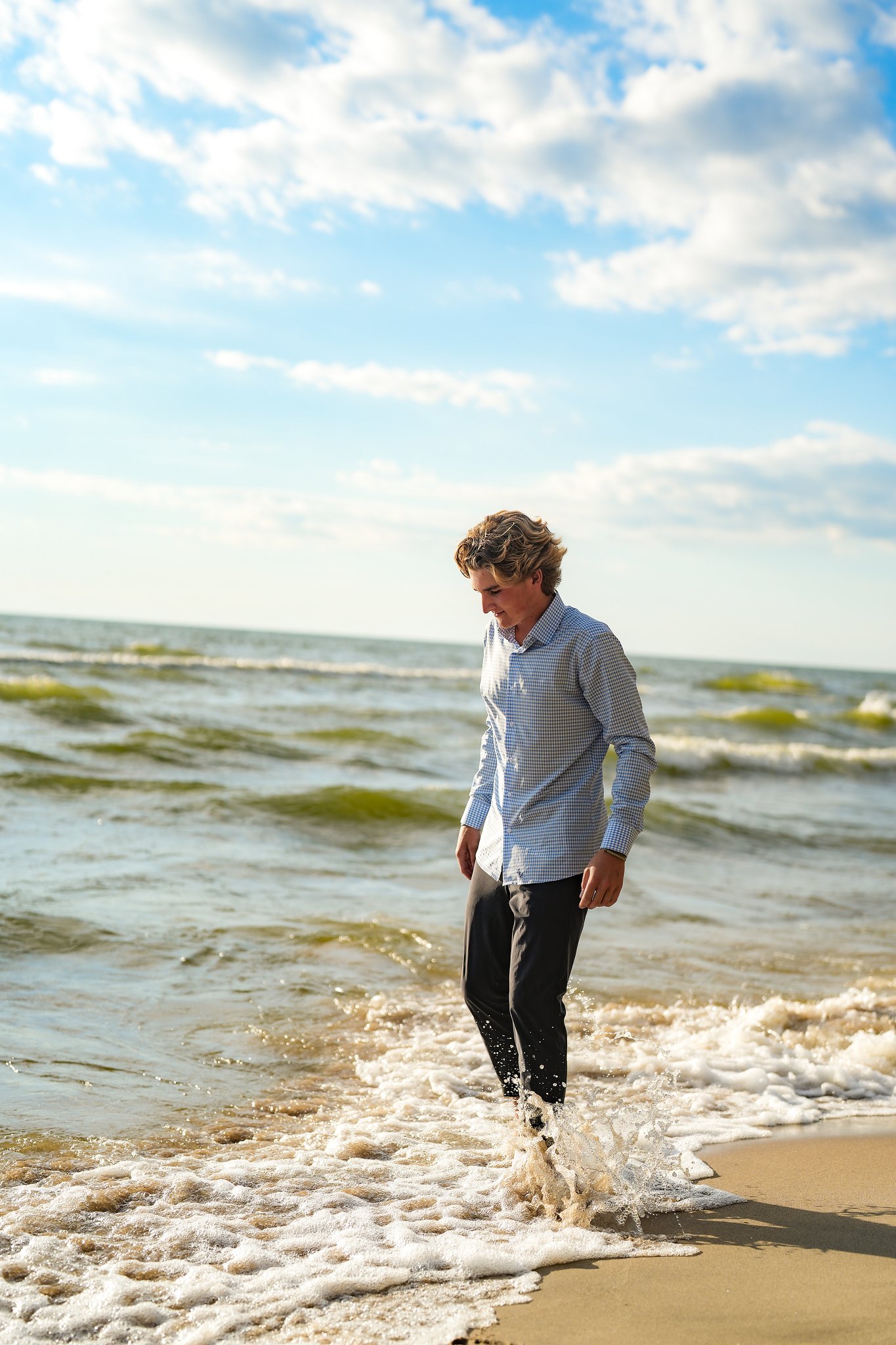 A young man with curly hair wearing a blue checkered shirt and black pants walking along the shoreline of the ocean with waves crashing at his feet under a partly cloudy sky.
