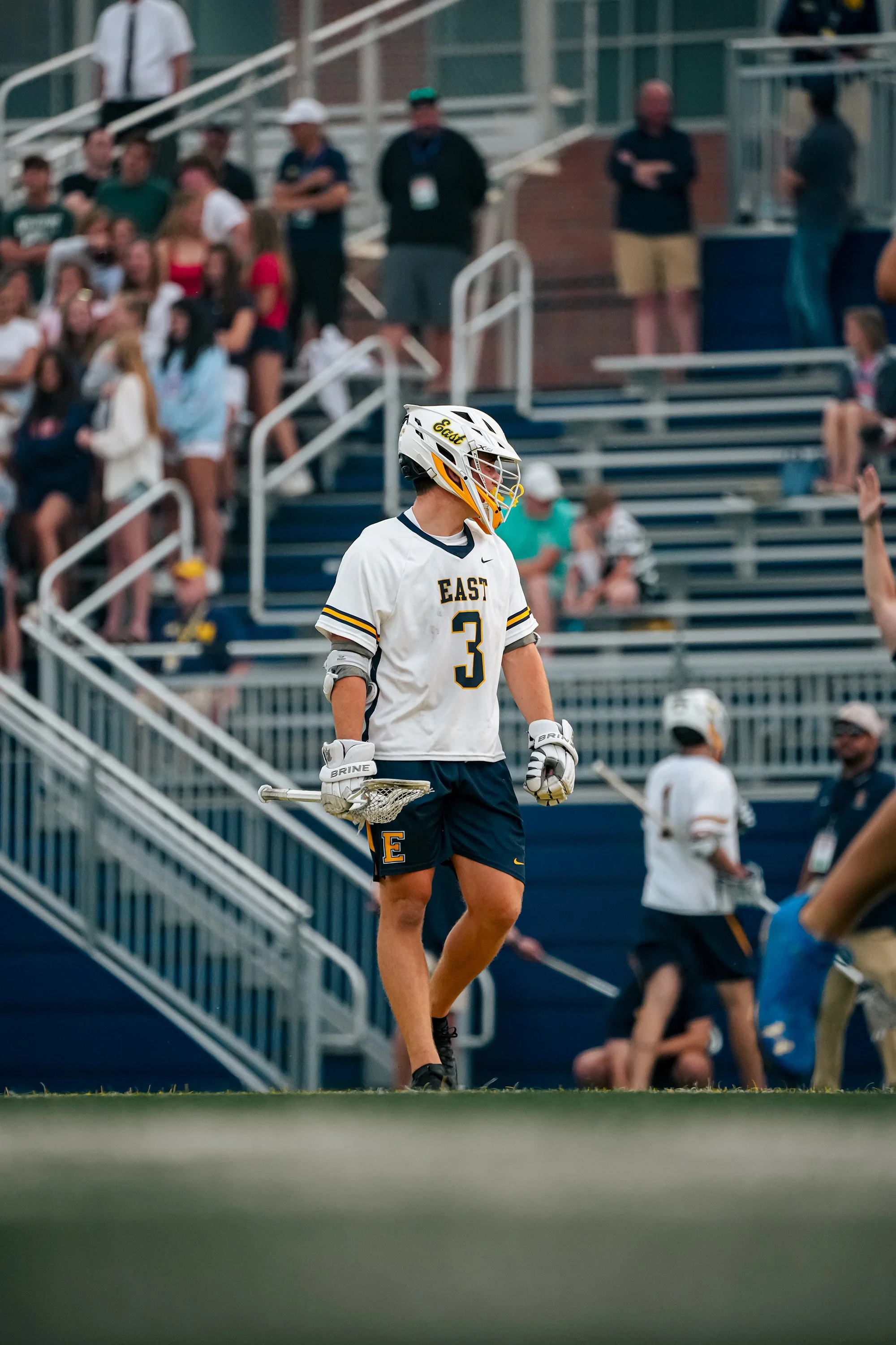 A lacrosse player in a white jersey with 'EAST' and the number 3, holding a lacrosse stick, walking across a sports field with spectators and people in the background.