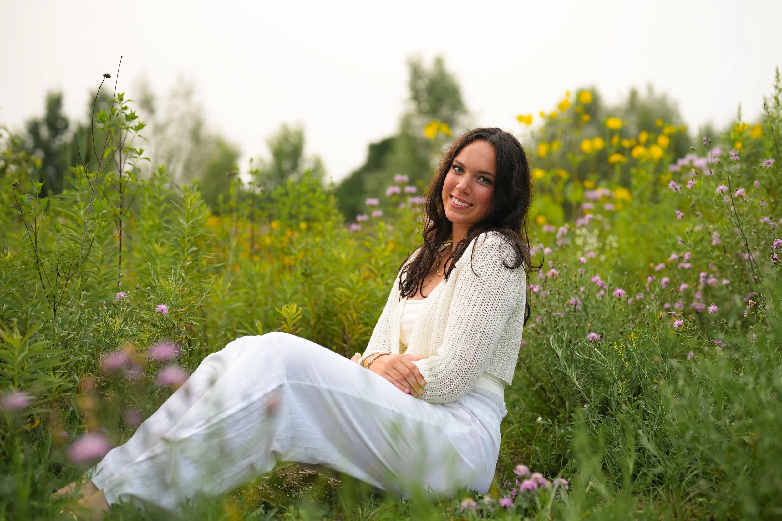 A woman with dark hair, wearing a white cardigan and white skirt, sitting in a flower field with pink, yellow, and purple blossoms and trees in the background, smiling at the camera.