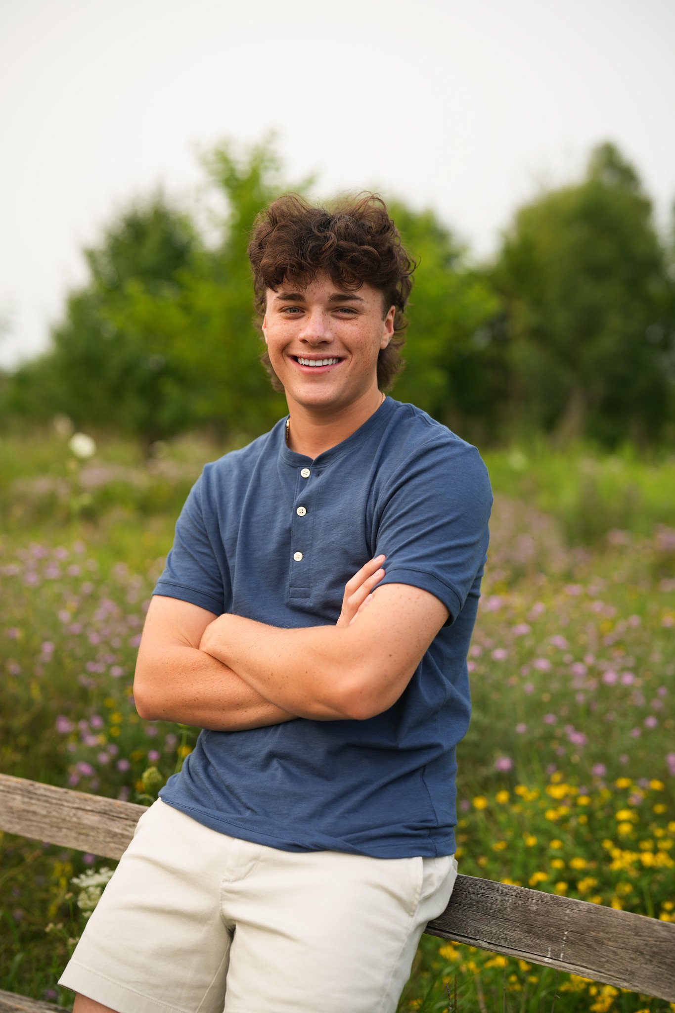 A young man with curly brown hair, wearing a navy blue polo shirt and beige shorts, stands outdoors in a field of purple and yellow flowers with green trees in the background, smiling and crossing his arms.
