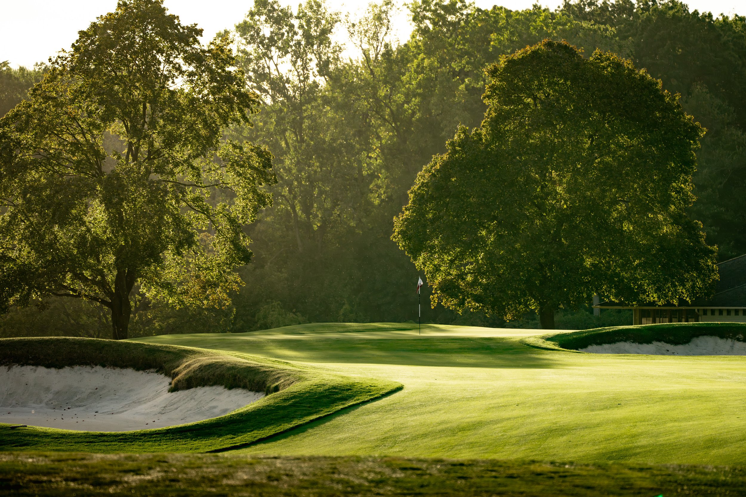 A golf course with lush green grass, a sand trap on the left, and a flagstick on the green surrounded by trees, with sunlight filtering through the trees.