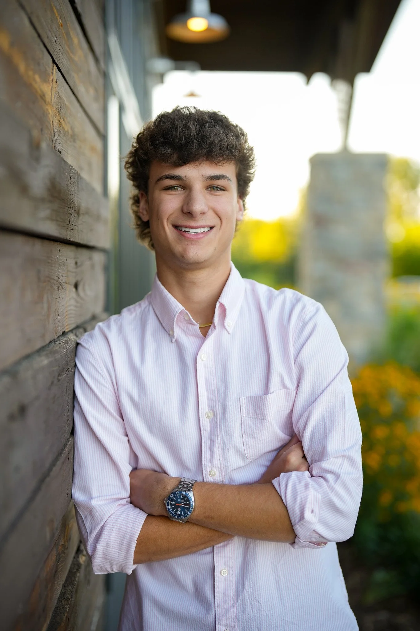 Young man with curly hair smiling, standing outdoors by a wooden wall with arms crossed, wearing a light pink striped shirt and a watch, with greenery and blurred background.