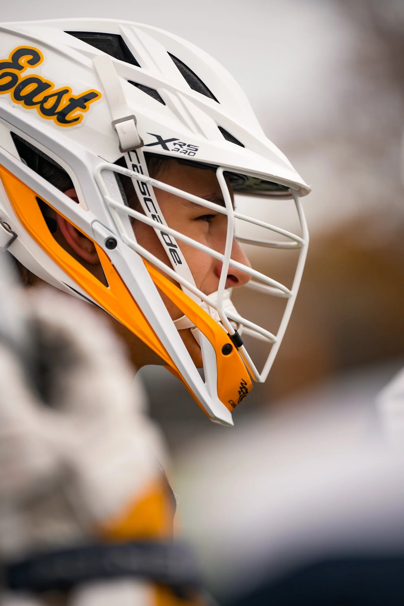 Close-up of a person wearing a white ice hockey goalie helmet with orange and black accents, and a cage face mask, focusing on the side and eye area.