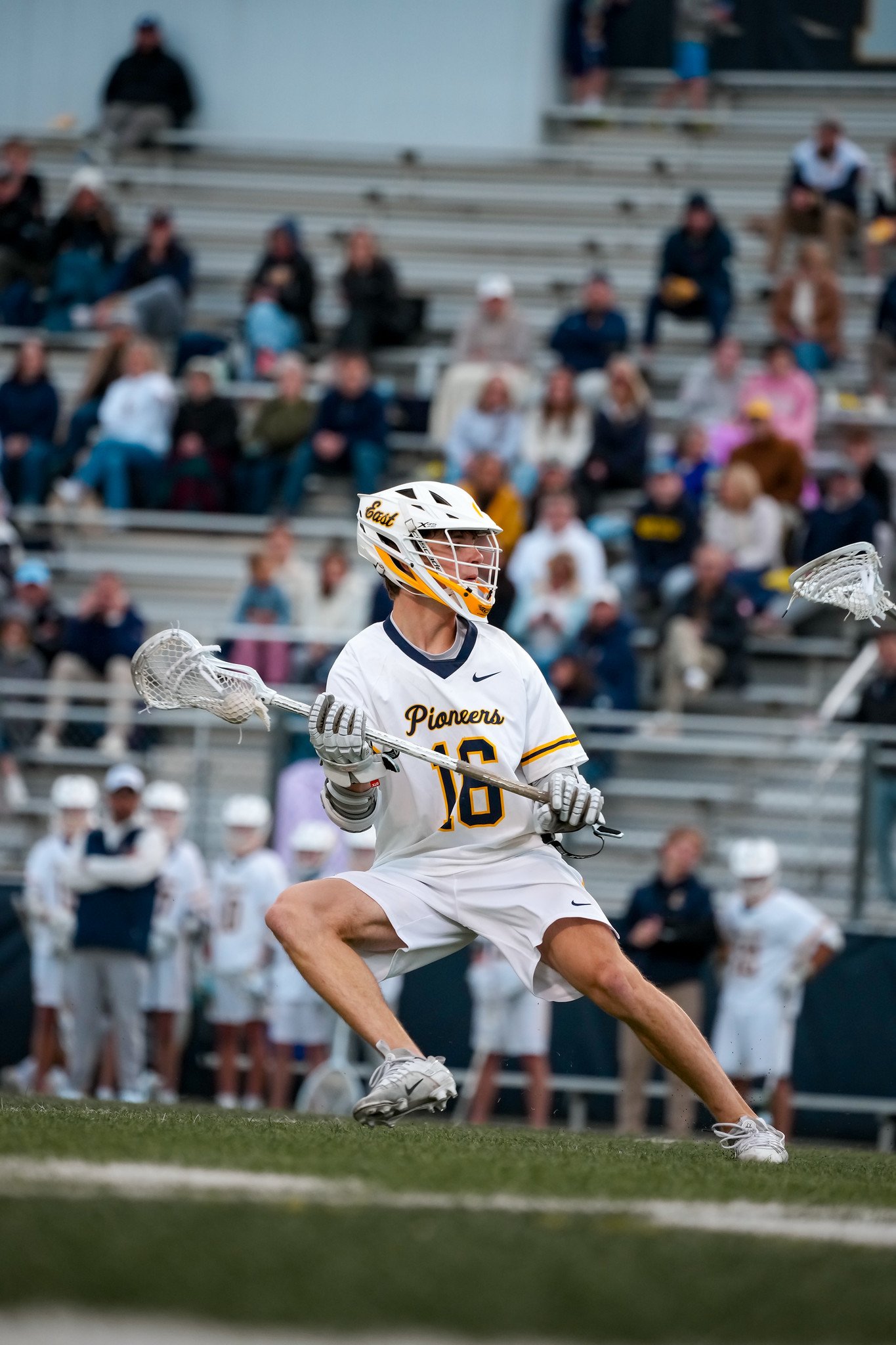 Lacrosse player in white jersey with number 16, wearing a helmet labeled East, holding a lacrosse stick, engaged in a game on a grassy field with spectators in the bleachers.