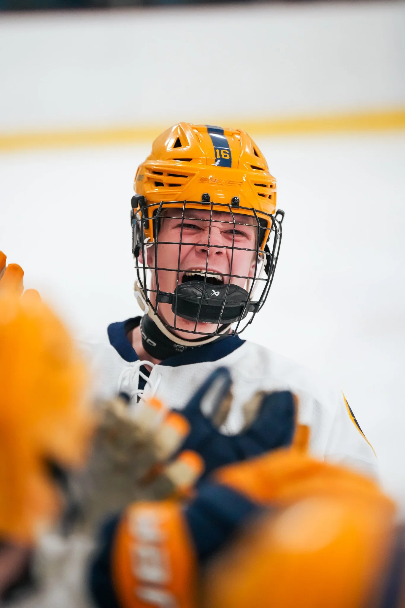 Close-up of a young ice hockey player wearing a yellow helmet, black cage face mask, and gloves, celebrating on the ice rink with a joyful expression.