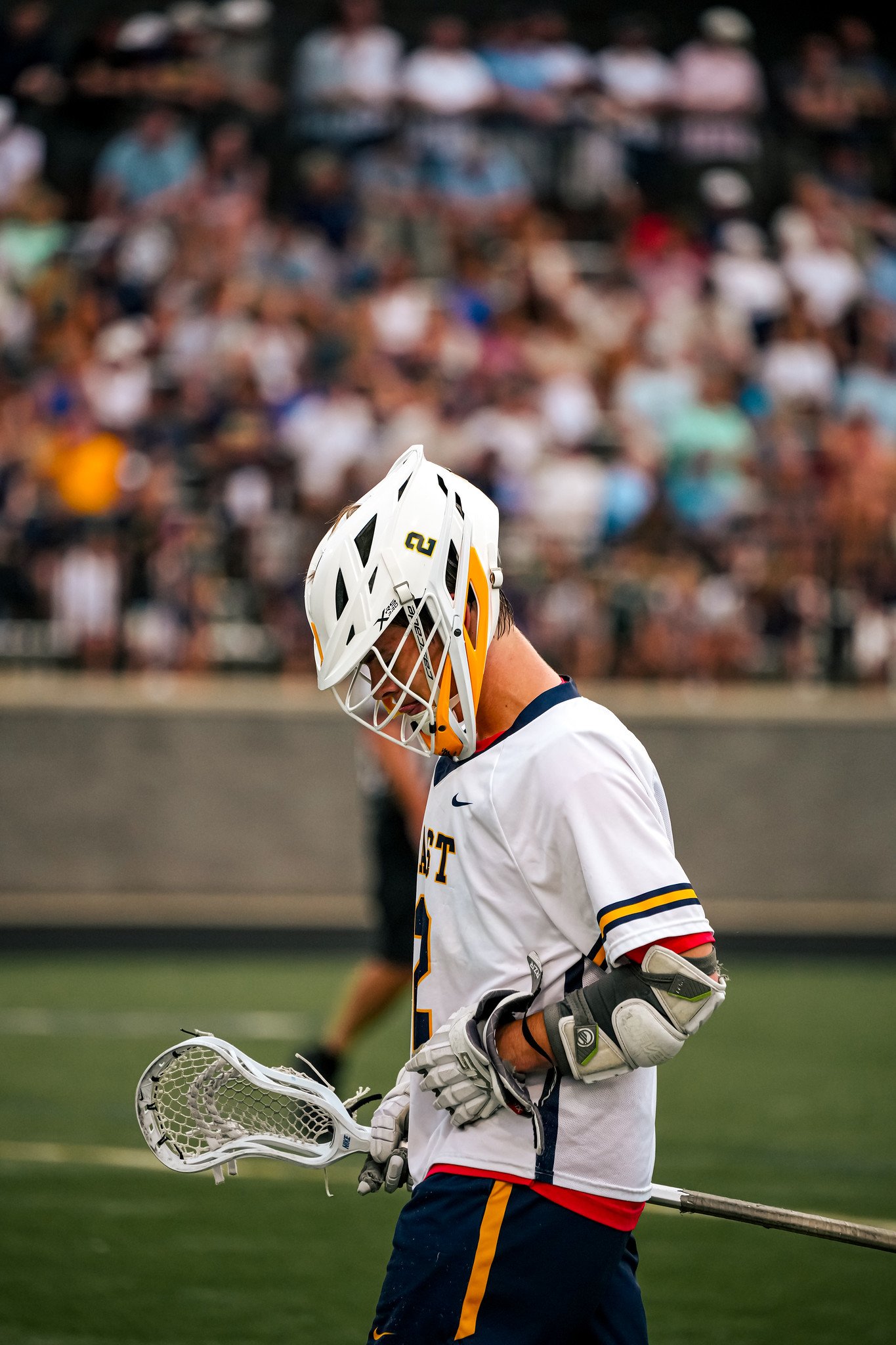 Lacrosse player in uniform and helmet holding lacrosse stick, with a blurred crowd in the background.