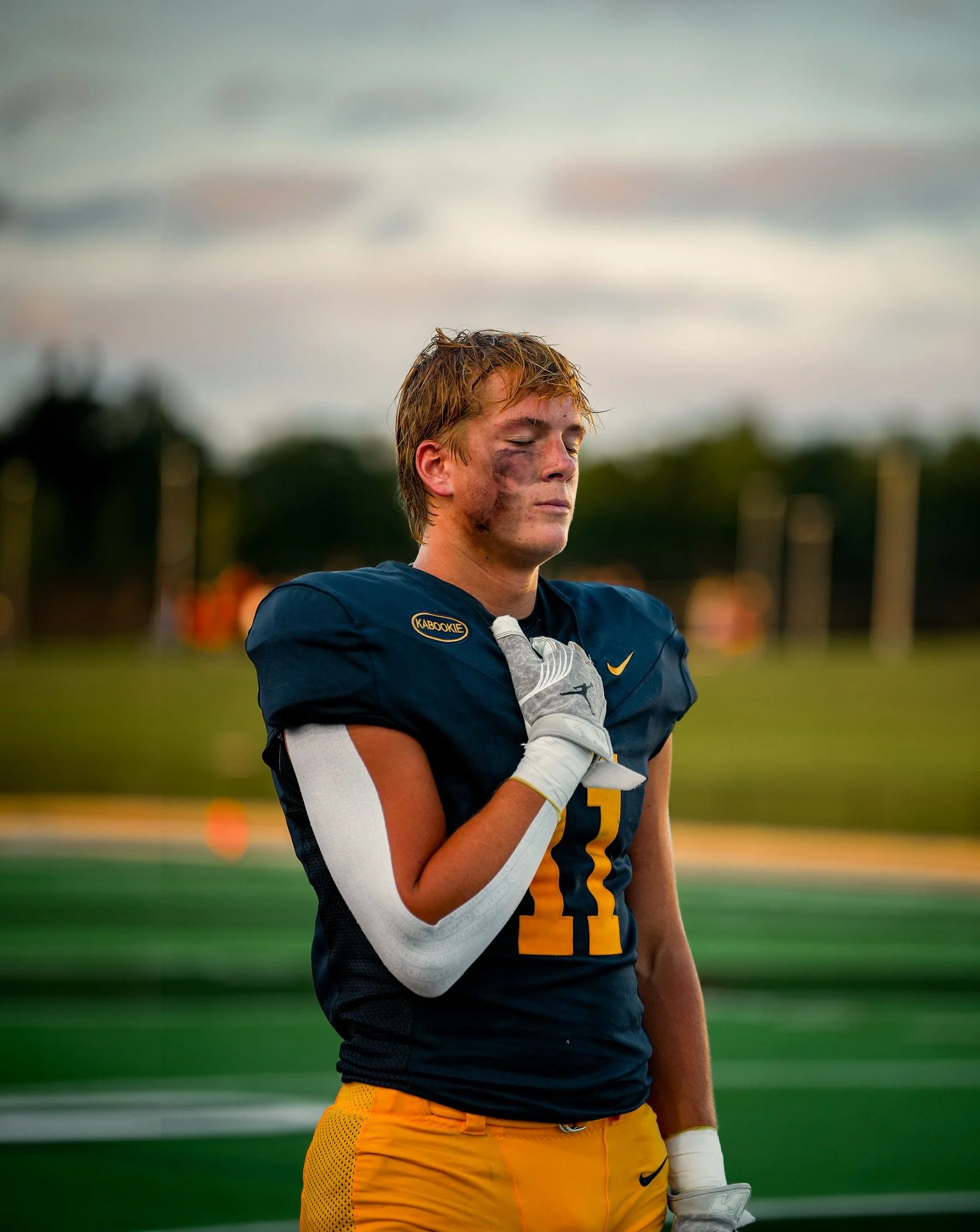 A football player with long red hair, wearing a navy blue jersey with yellow accents and the number 12, standing on the field during a game, with his eyes closed and one hand over his heart. The player has dirt on his face and is wearing a white glov