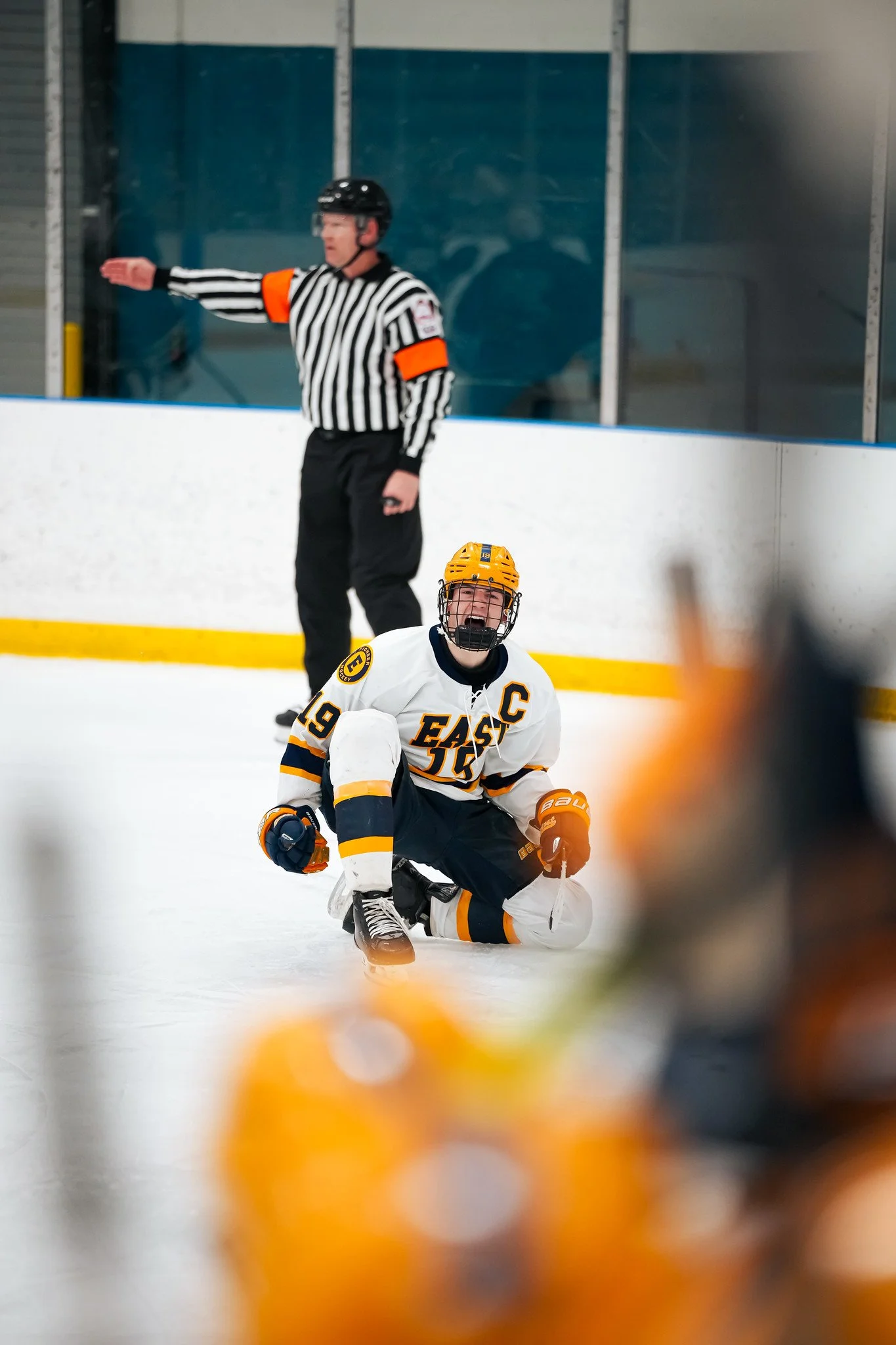 An ice hockey player wearing a white jersey with the number 19 and a captain's 'C' on the front, kneeling on the ice with an intense expression, and a referee in a black and white striped shirt and black helmet standing in the background near the rin