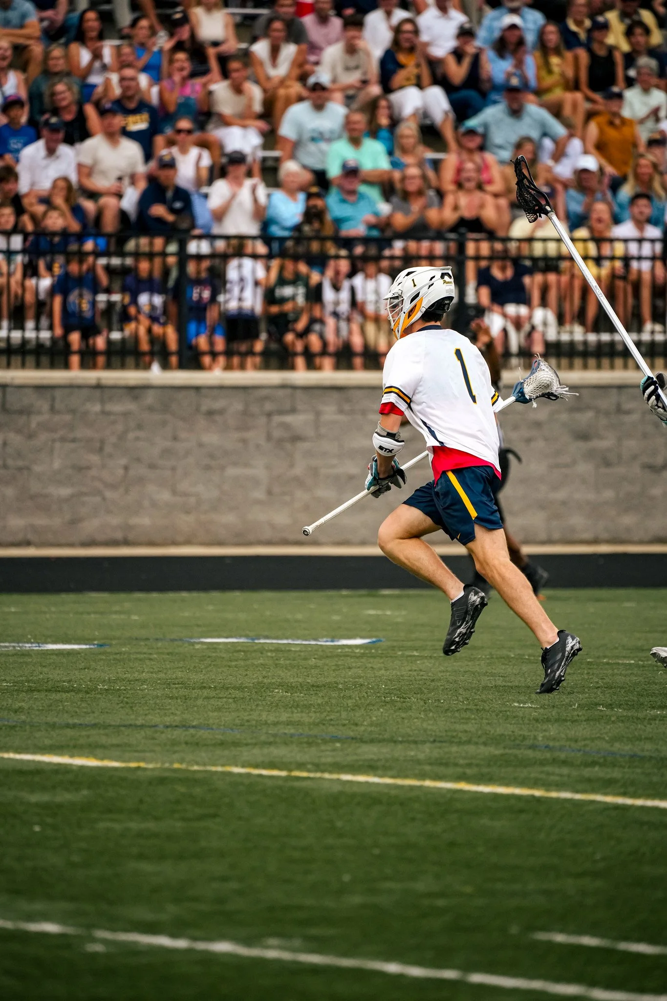 A lacrosse player in a white jersey with the number 1 and red, blue, and yellow accents runs across a green turf field during a game, holding a lacrosse stick. Spectators are seated behind a black fence, watching the game.