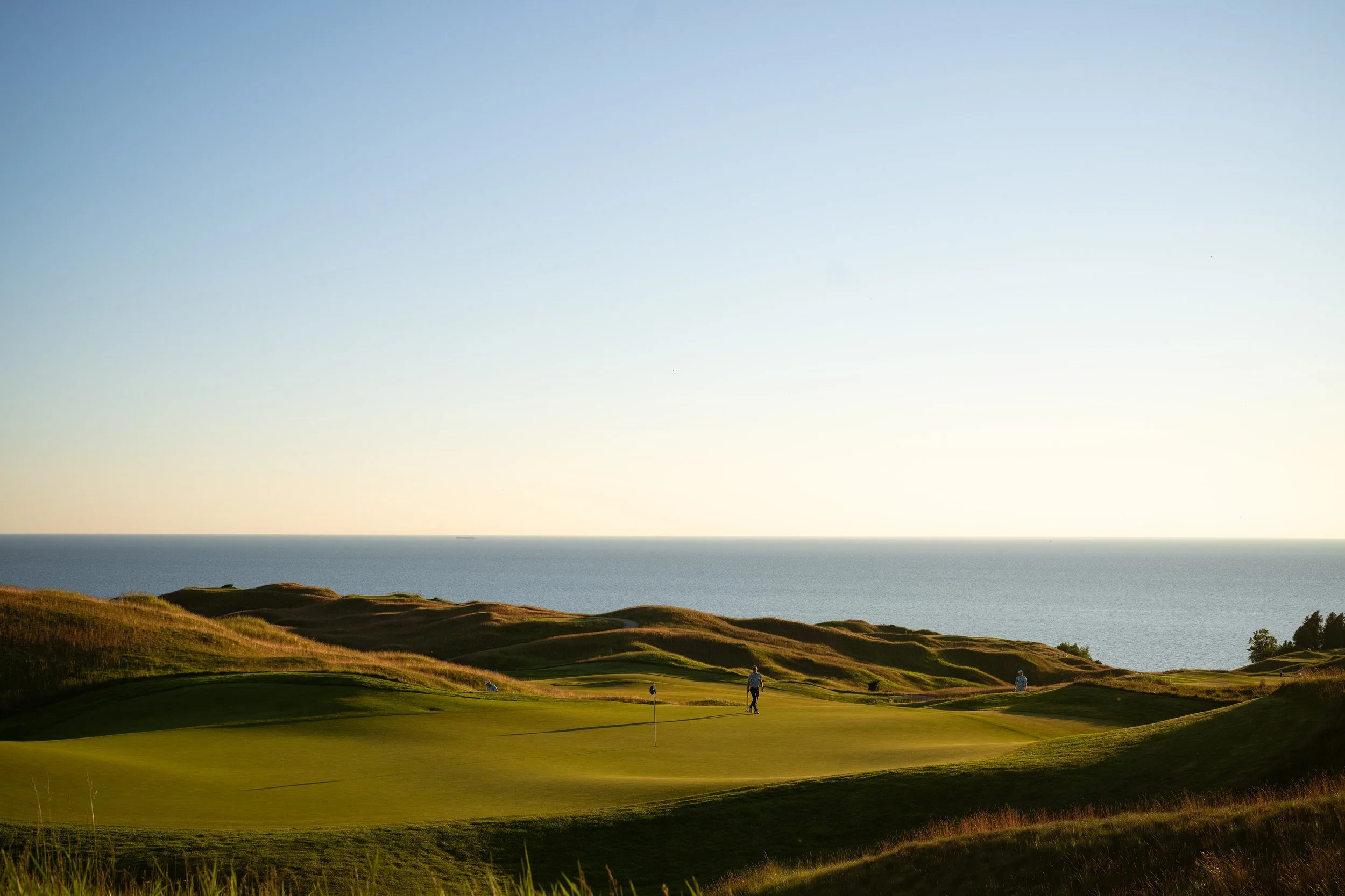 Golf course with green fairways and a flagstick, overlooking the ocean with hills in the foreground under a clear sky.
