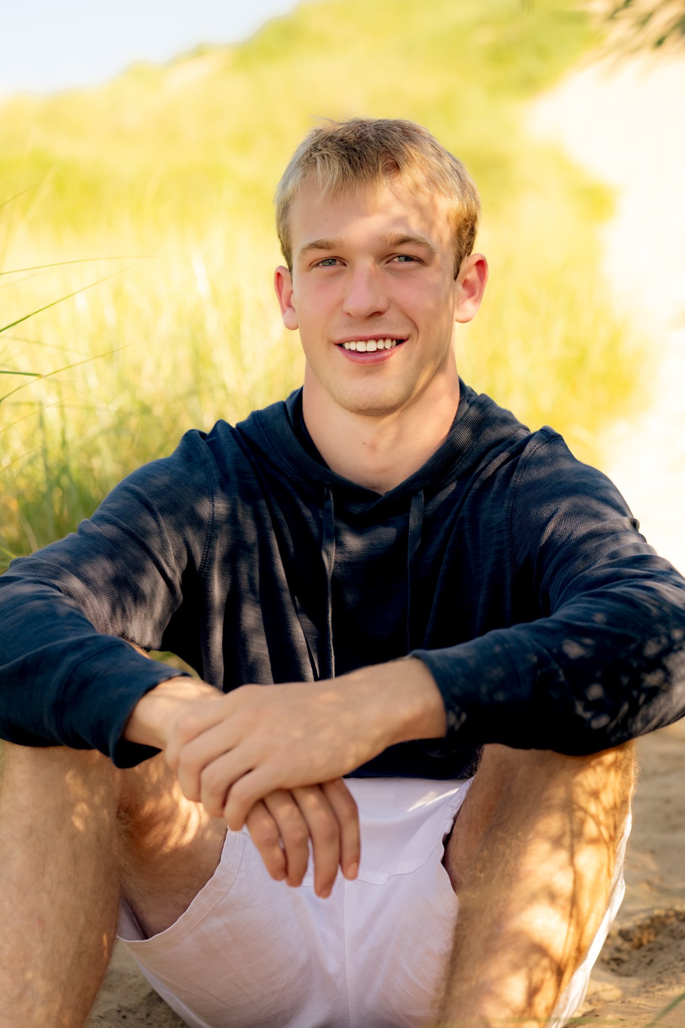 A young man with short blond hair smiling outdoors in a sunny, natural setting, with grass and trees in the background.