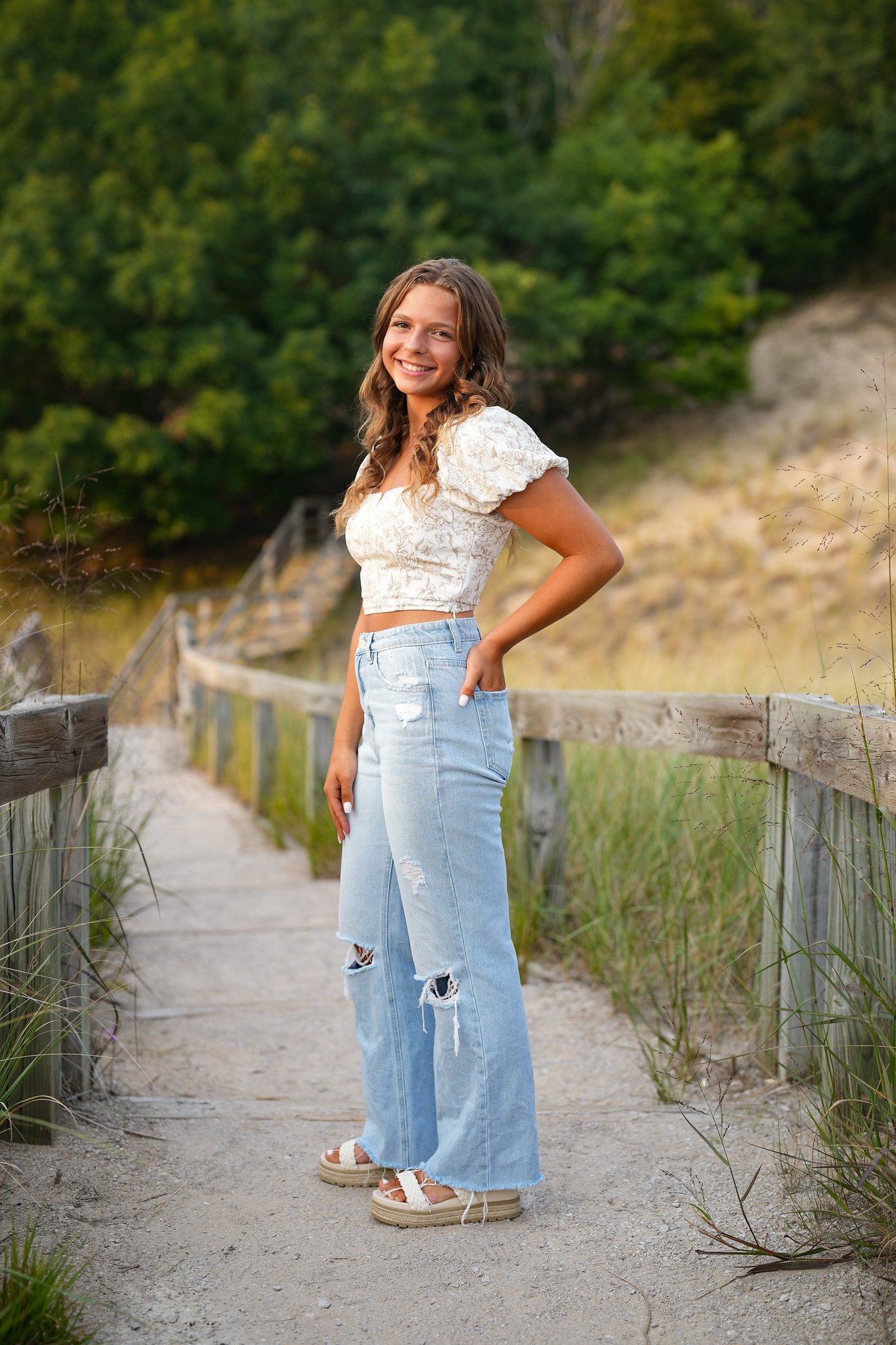 Young woman smiling on a sandy path near a wooden fence with greenery in the background, wearing a white cropped top, ripped jeans, and platform sandals.