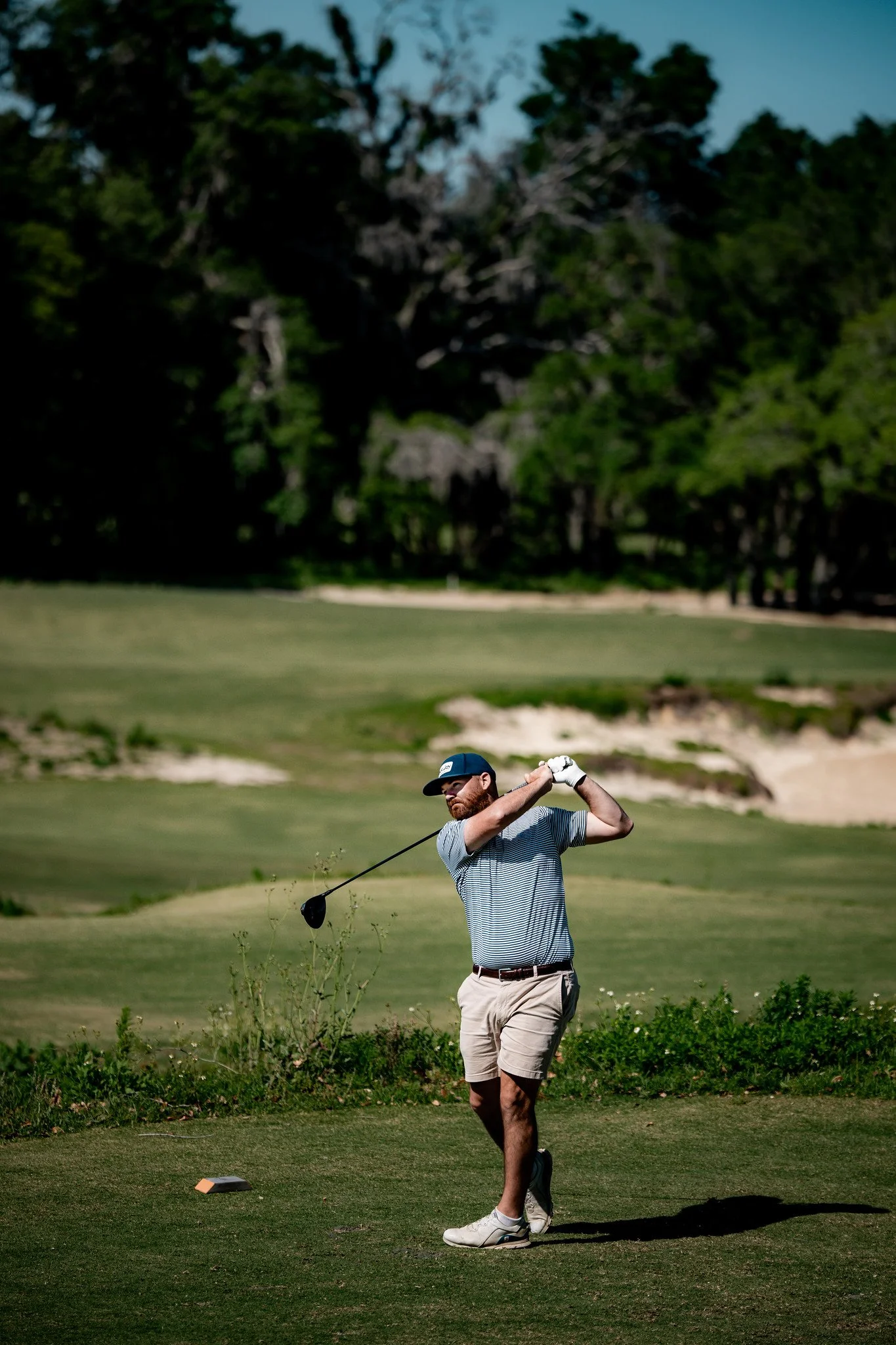 A man playing golf on a course, swinging a golf club, wearing a striped shirt, khaki shorts, and a cap, with a green landscape and trees in the background.