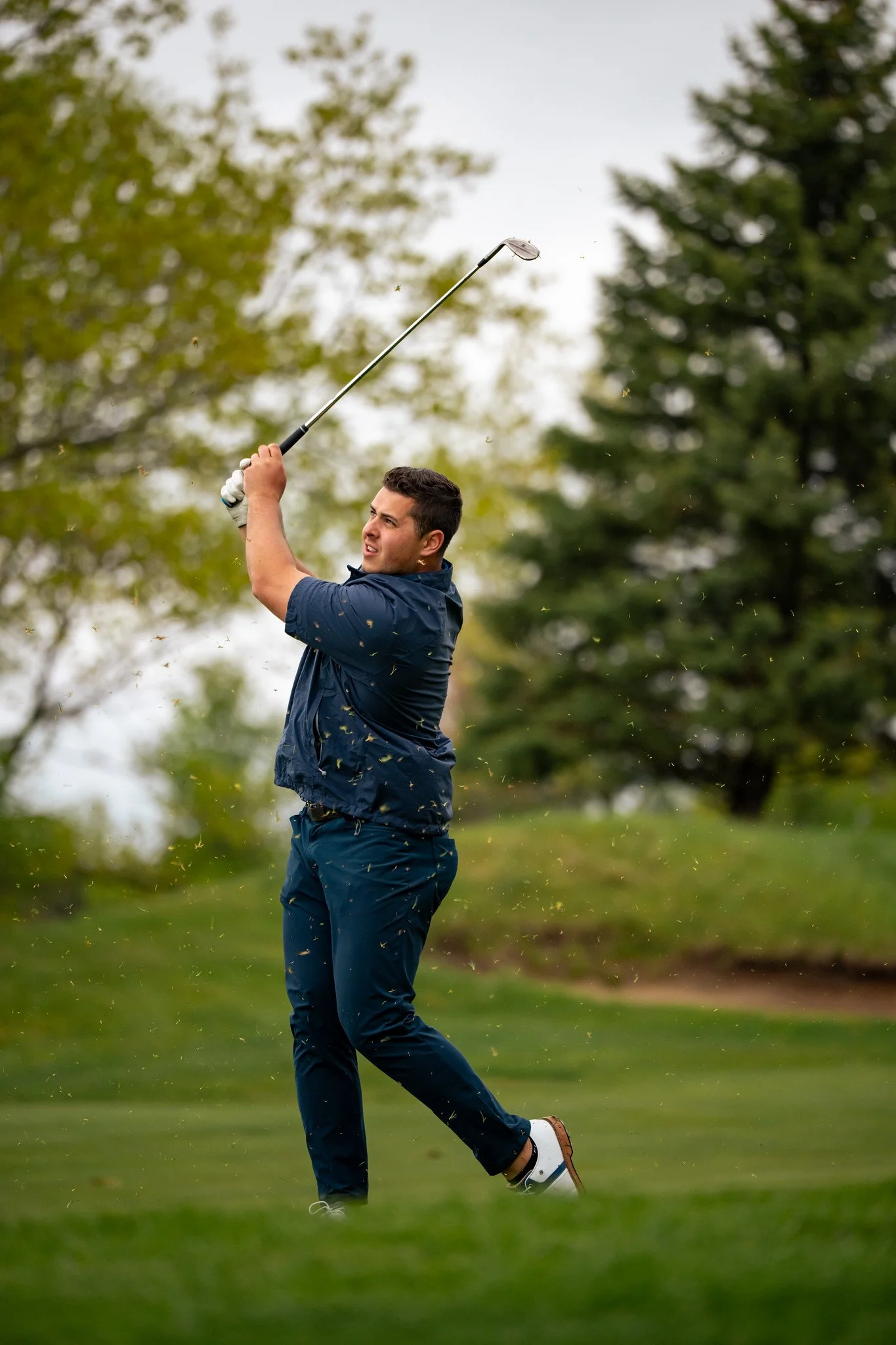 A man playing golf on a green field with trees in the background, swinging a golf club.