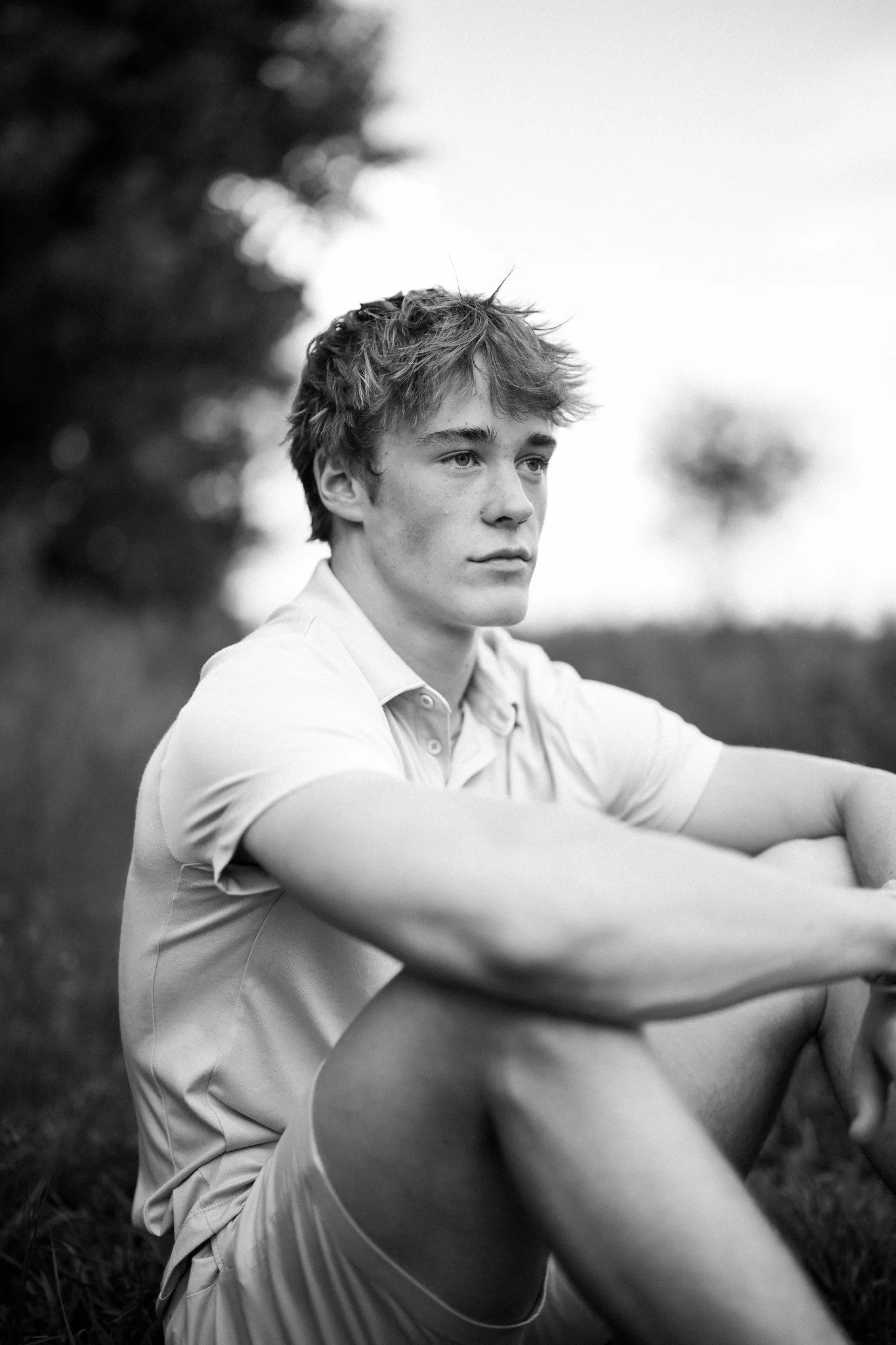 Black and white photo of a young man sitting outdoors with his arm resting on his knee, looking thoughtfully into the distance.