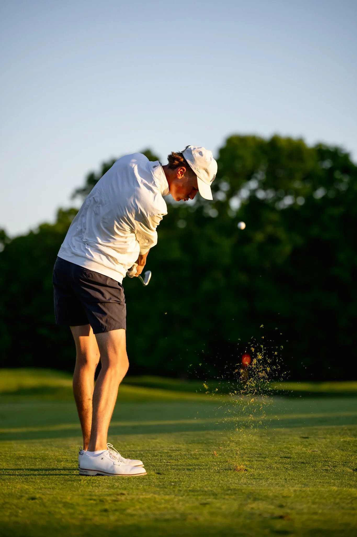 A man playing golf on a green field during sunset, wearing a white cap, white jacket, black shorts, and white golf shoes, taking a shot at the golf ball on the ground.
