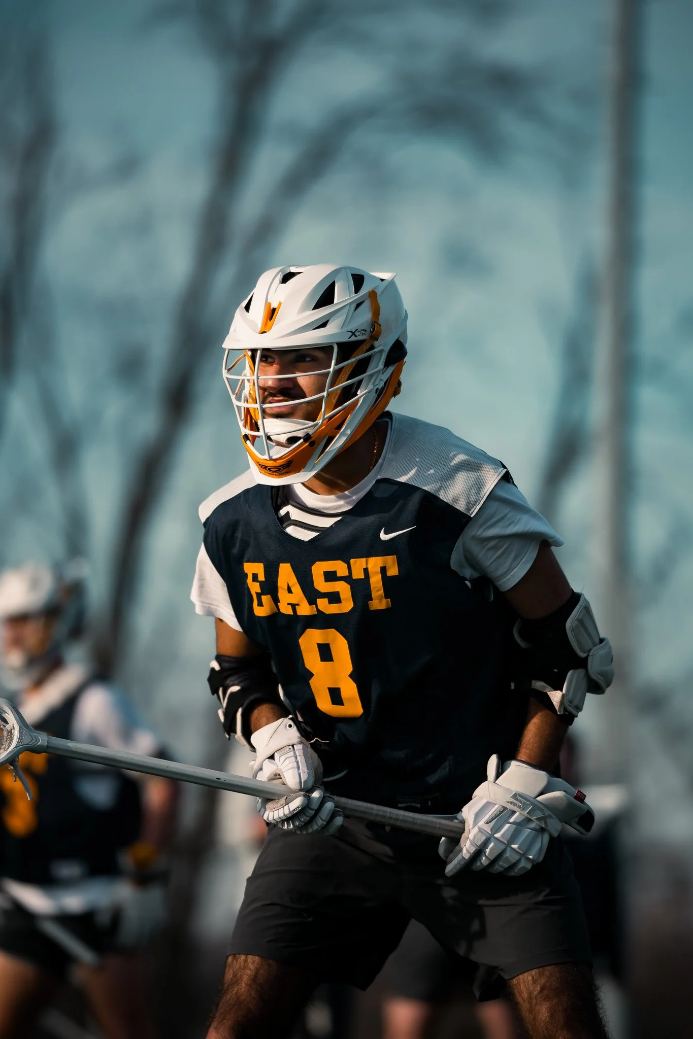 A male lacrosse player wearing a white helmet, black and white jersey with the word 'EAST' and the number 8, and gloves, holding a lacrosse stick during a game outdoor in a park with leafless trees in the background.