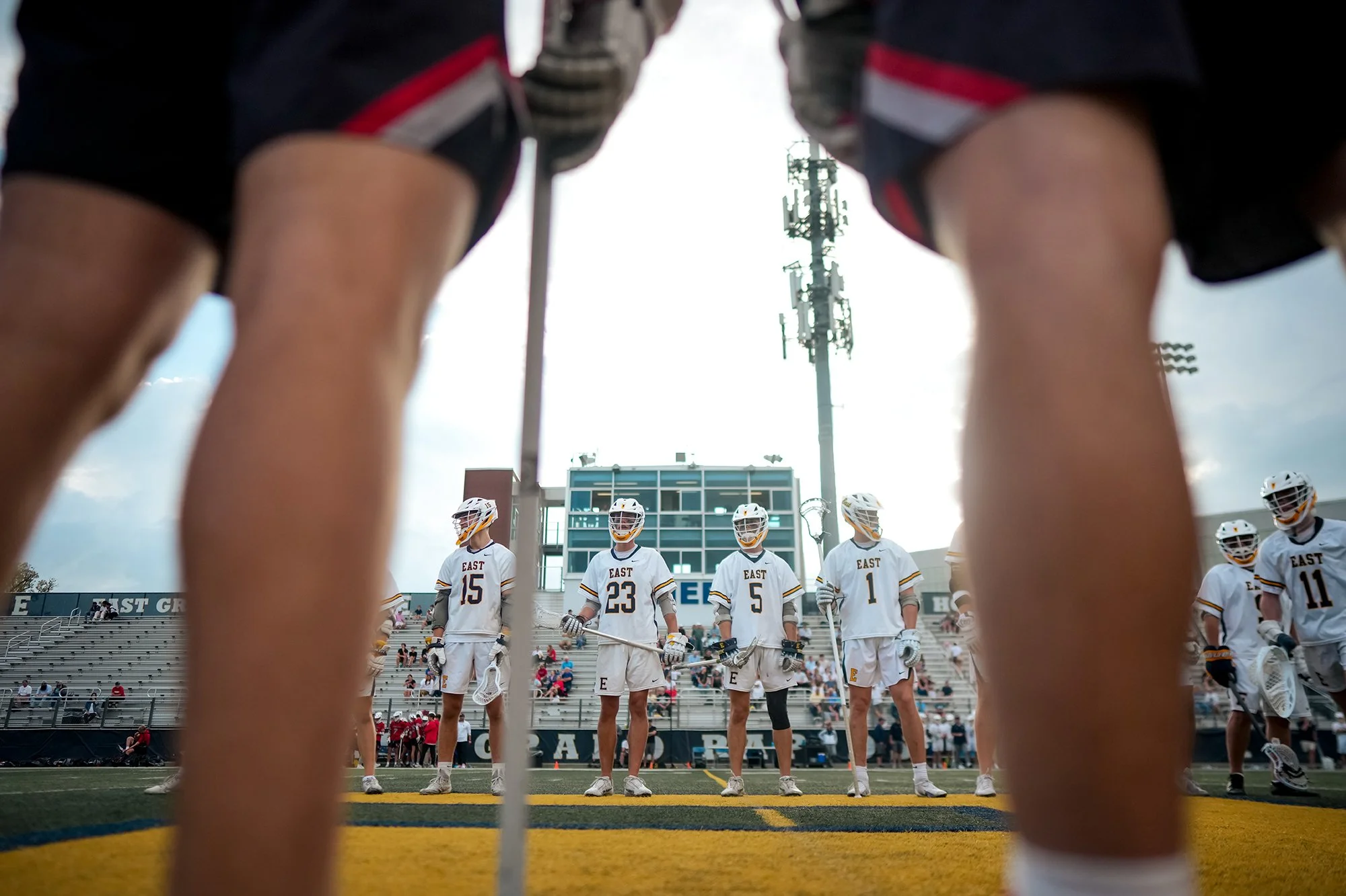 A group of lacrosse players standing on a field, viewed from beneath a lacrosse goal net, with spectators in the stands and a stadium building with bleachers and a tall transmission tower in the background.