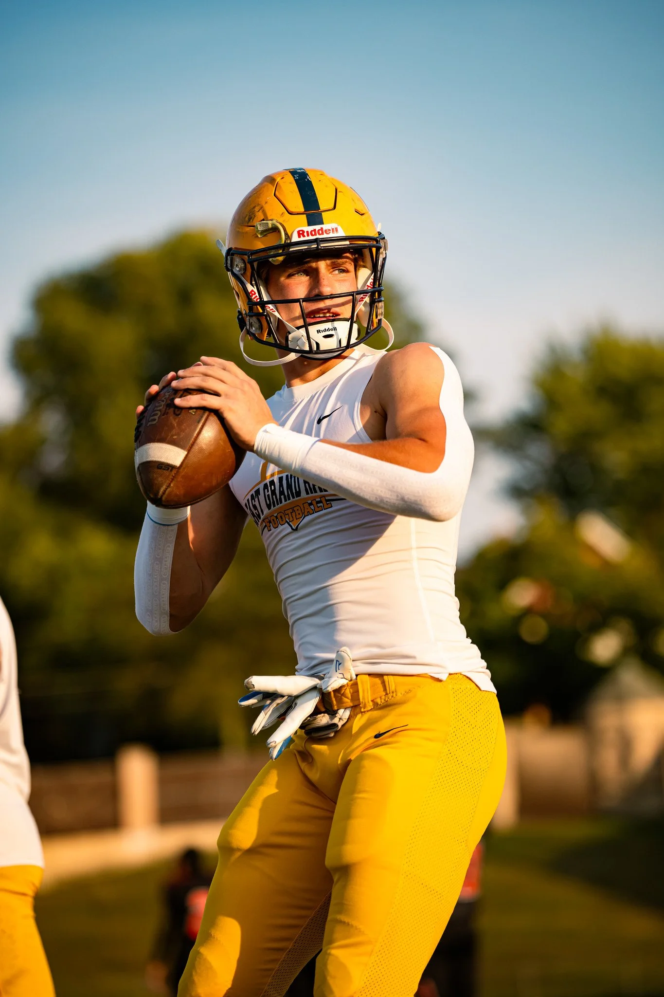 A football player wearing a yellow helmet and yellow pants holds a football, outdoors with trees and a clear sky in the background.