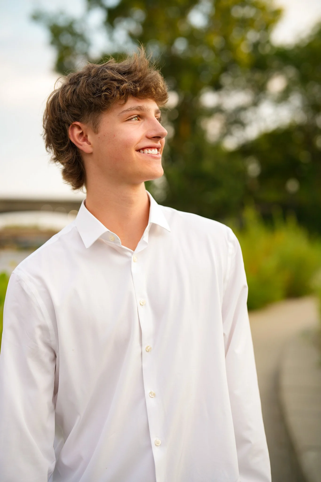 A young man with curly brown hair wearing a white shirt, smiling slightly, standing outdoors with blurred green trees in the background.