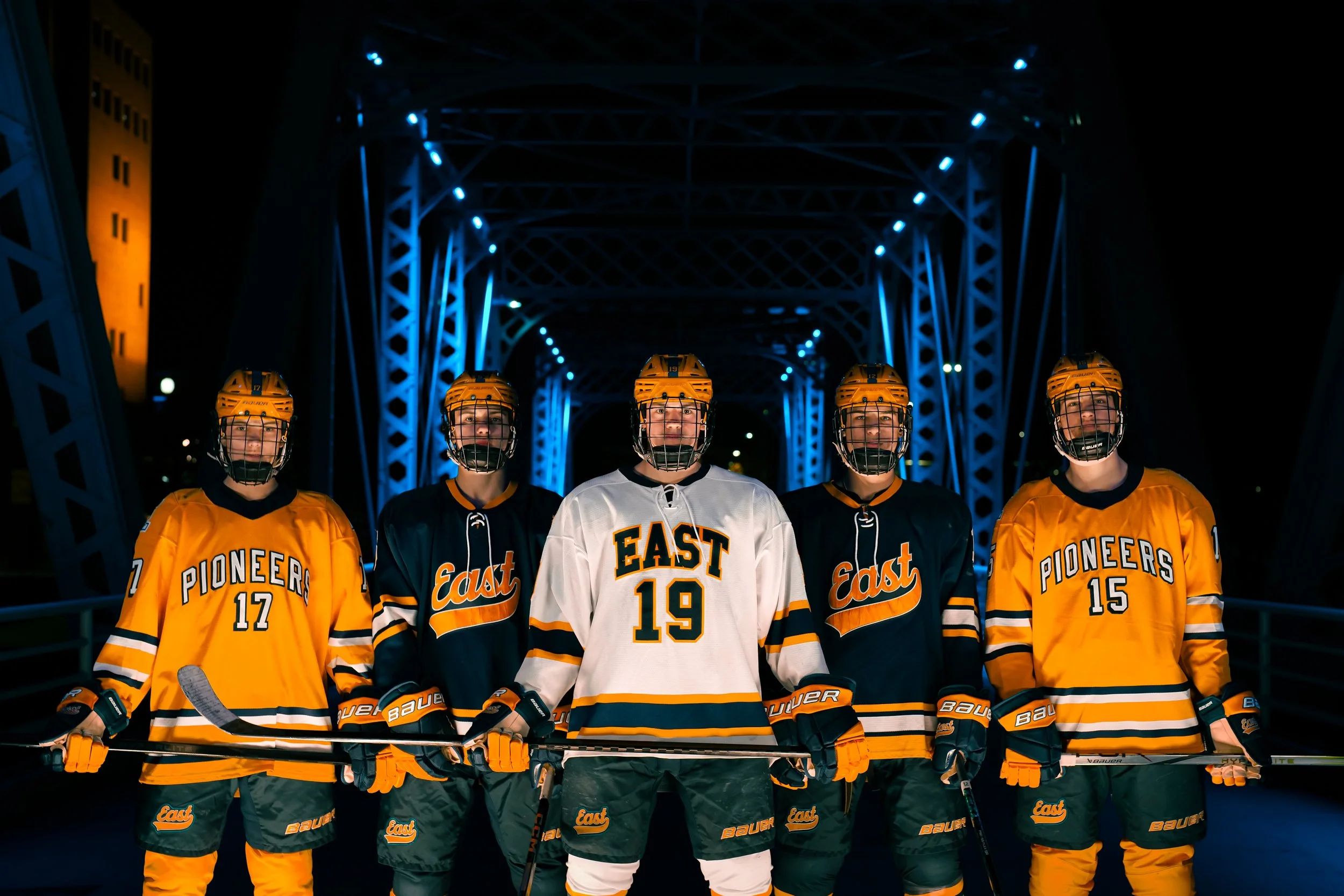 Five ice hockey players in yellow, black, and white uniforms standing on an ice rink under a bridge with blue lighting at night.